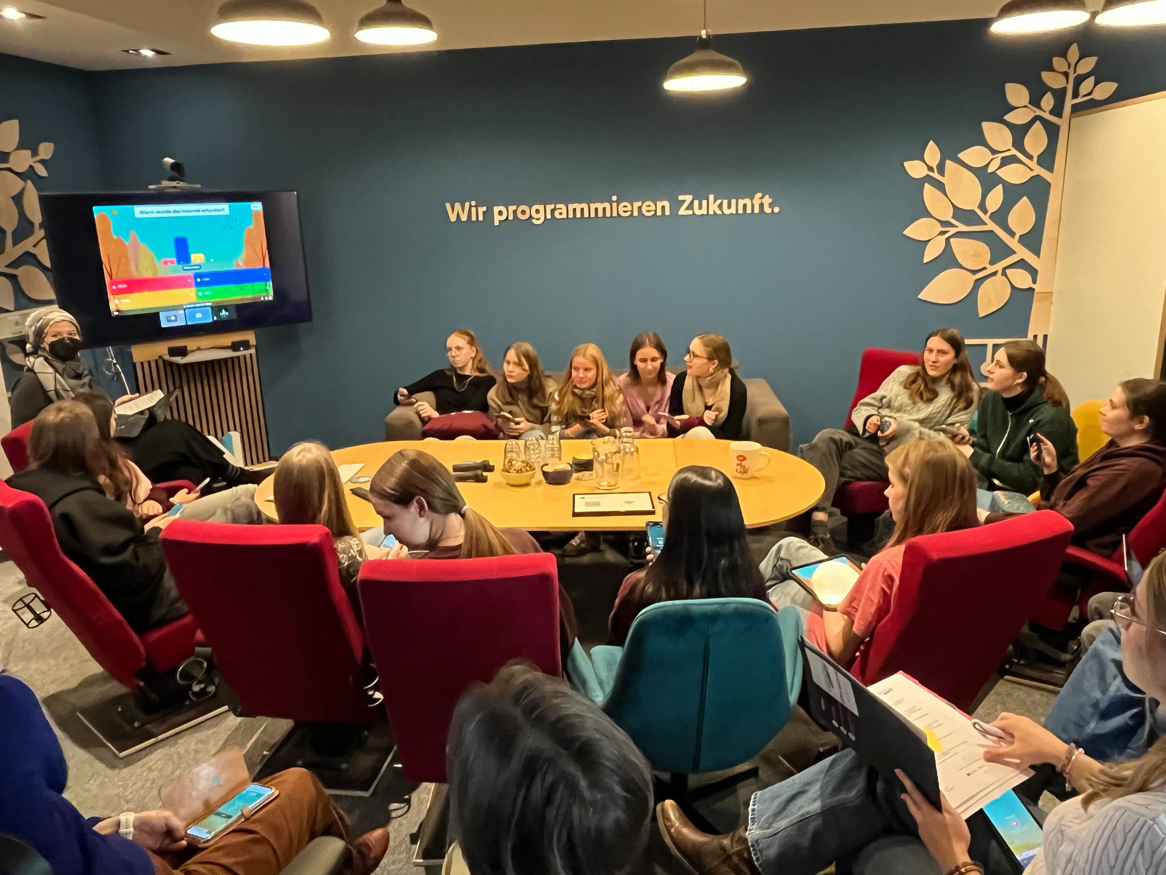 KI generiert: Eine Gruppe von Menschen sitzt in einem modernen Konferenzraum mit einem Bildschirm an der Wand, auf dem ein Spiel oder eine Präsentation zu sehen ist, während der Raumtext "Wir programmieren Zukunft" zeigt.