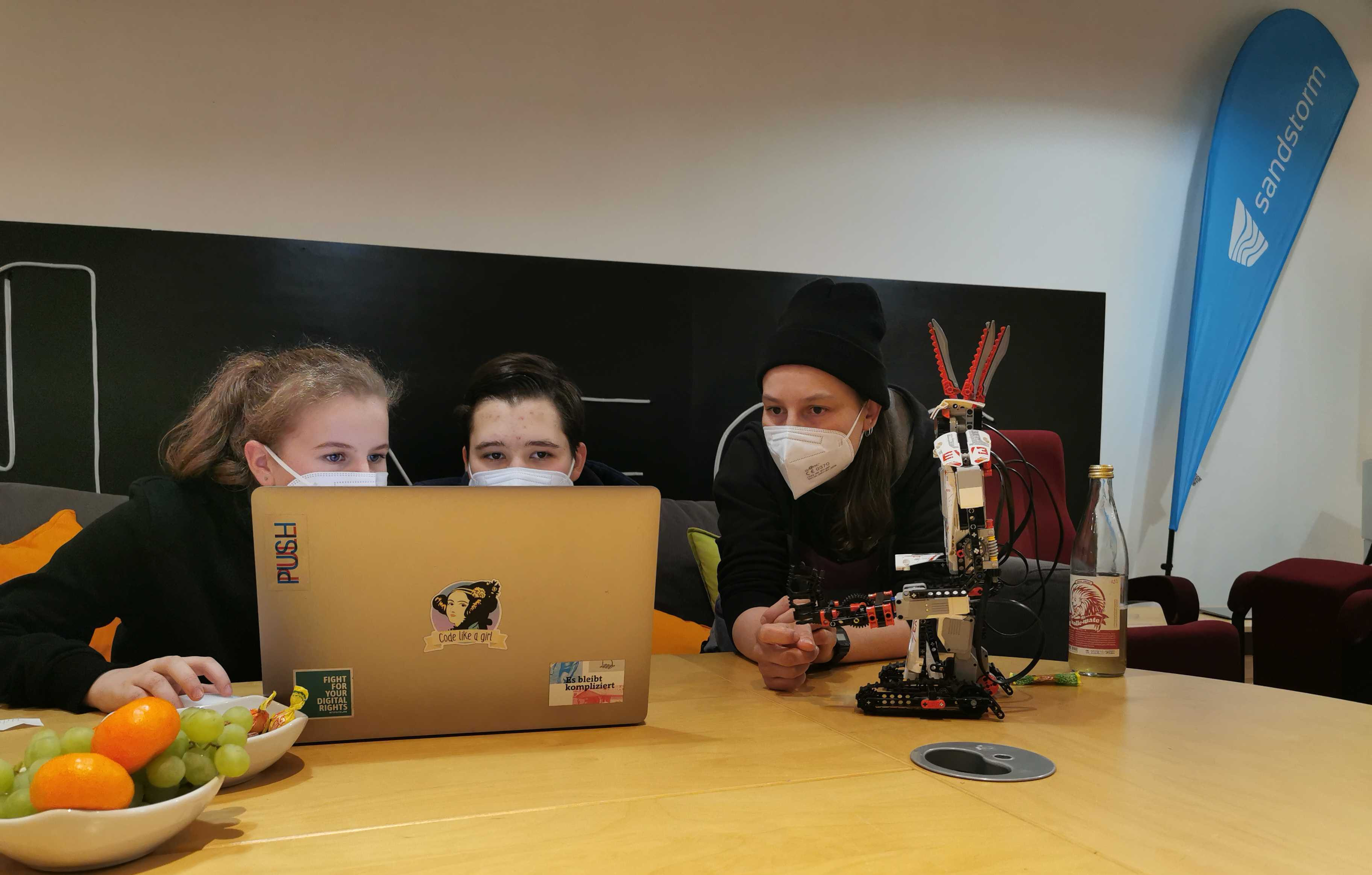 Zwei Schülerinnen und eine Programmiererin schauen auf einen Bildschirm, daneben steht ein Lego Mindstorms Roboter