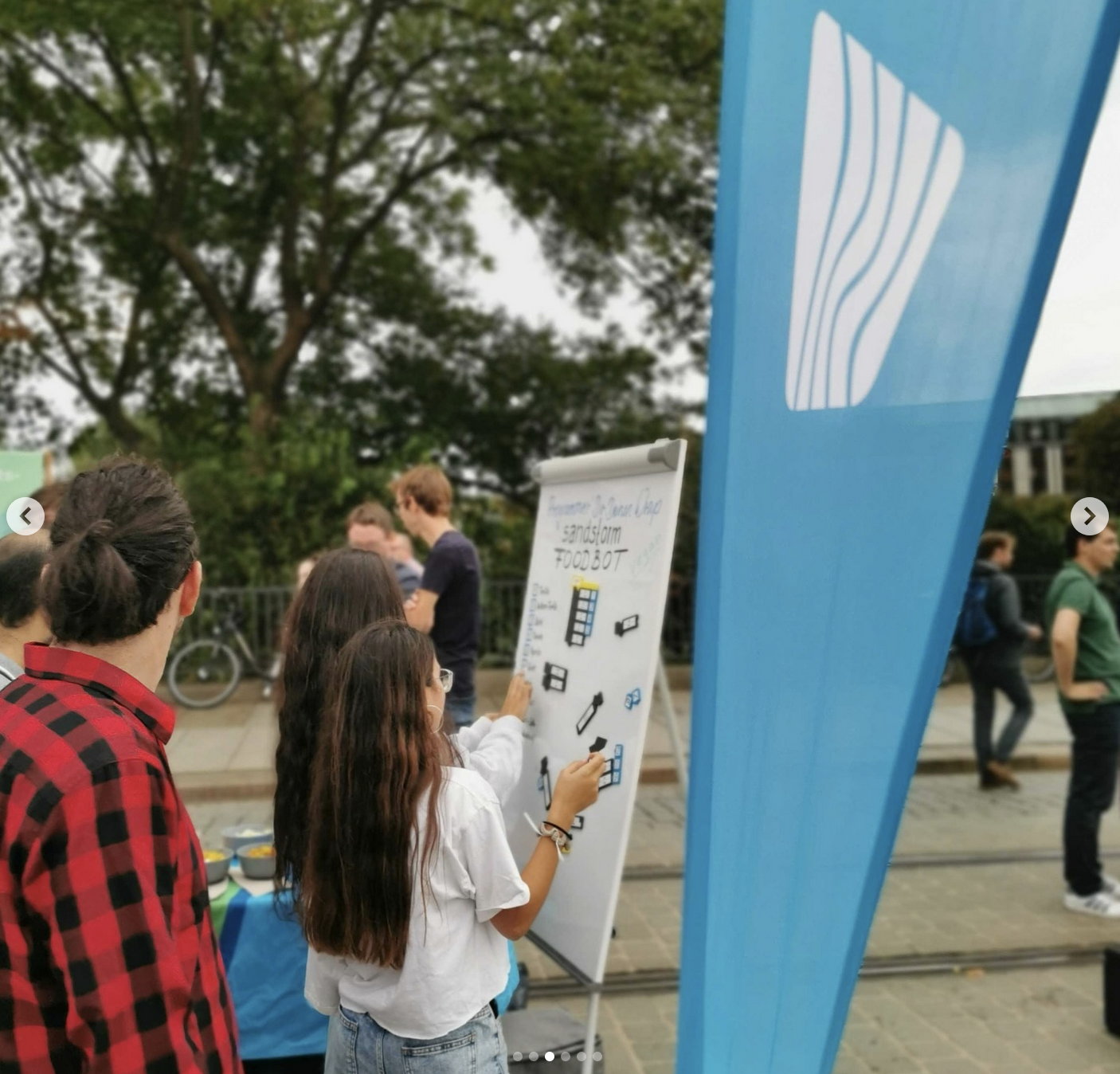 KI generiert: Das Bild zeigt mehrere Personen, die sich um ein Flipchart im Freien versammelt haben, um daran zu arbeiten oder es zu betrachten. Im Vordergrund ist eine blaue Fahne zu sehen.