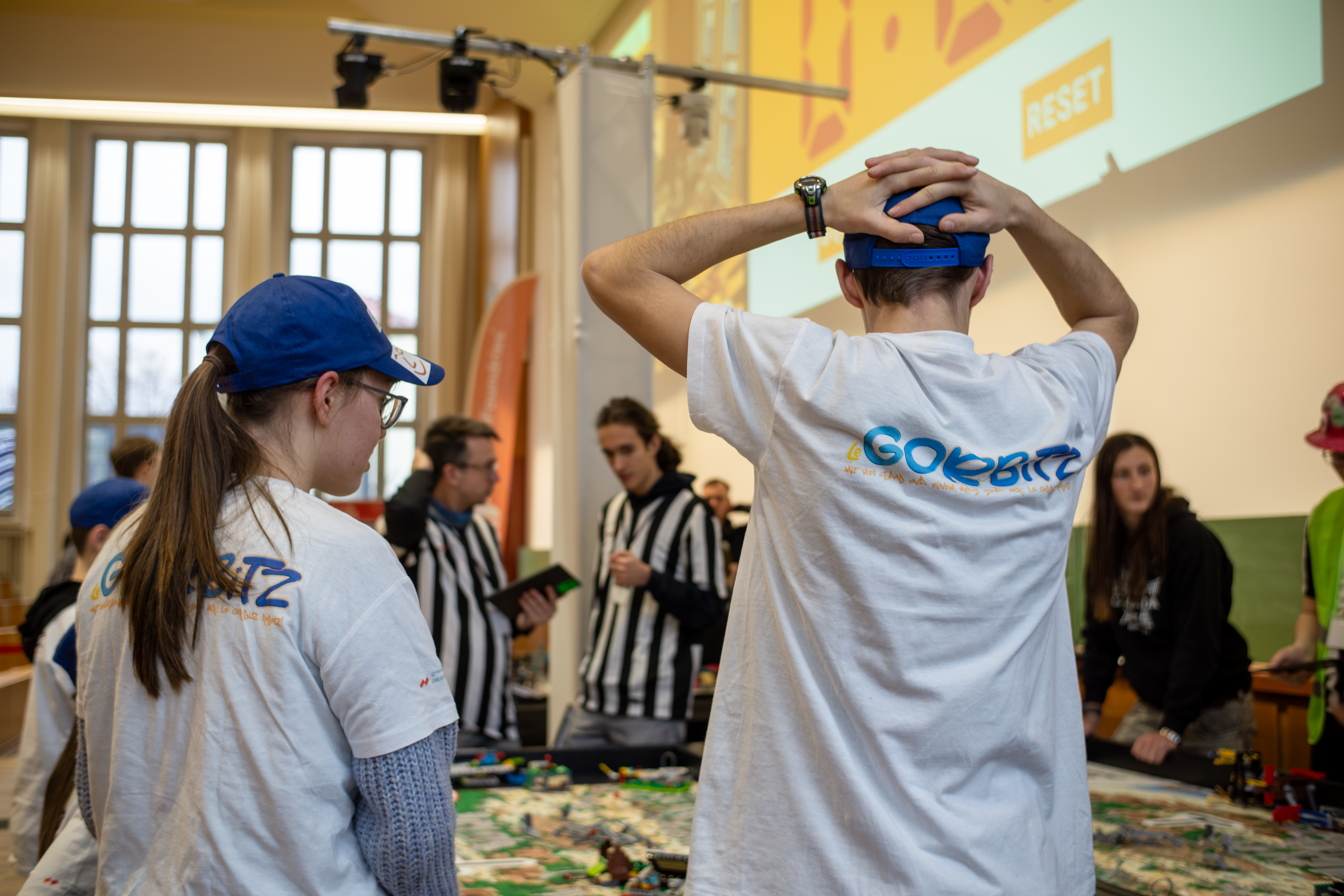 KI generiert: Das Bild zeigt eine Gruppe von Menschen, die in einem Raum mit großen Fenstern und einer Projektionswand an einem Tisch sitzen und sich auf ein Spiel oder eine Aktivität konzentrieren, wobei einige Teilnehmer die T-Shirts mit dem Aufdruck "GOLFBRZ" tragen.