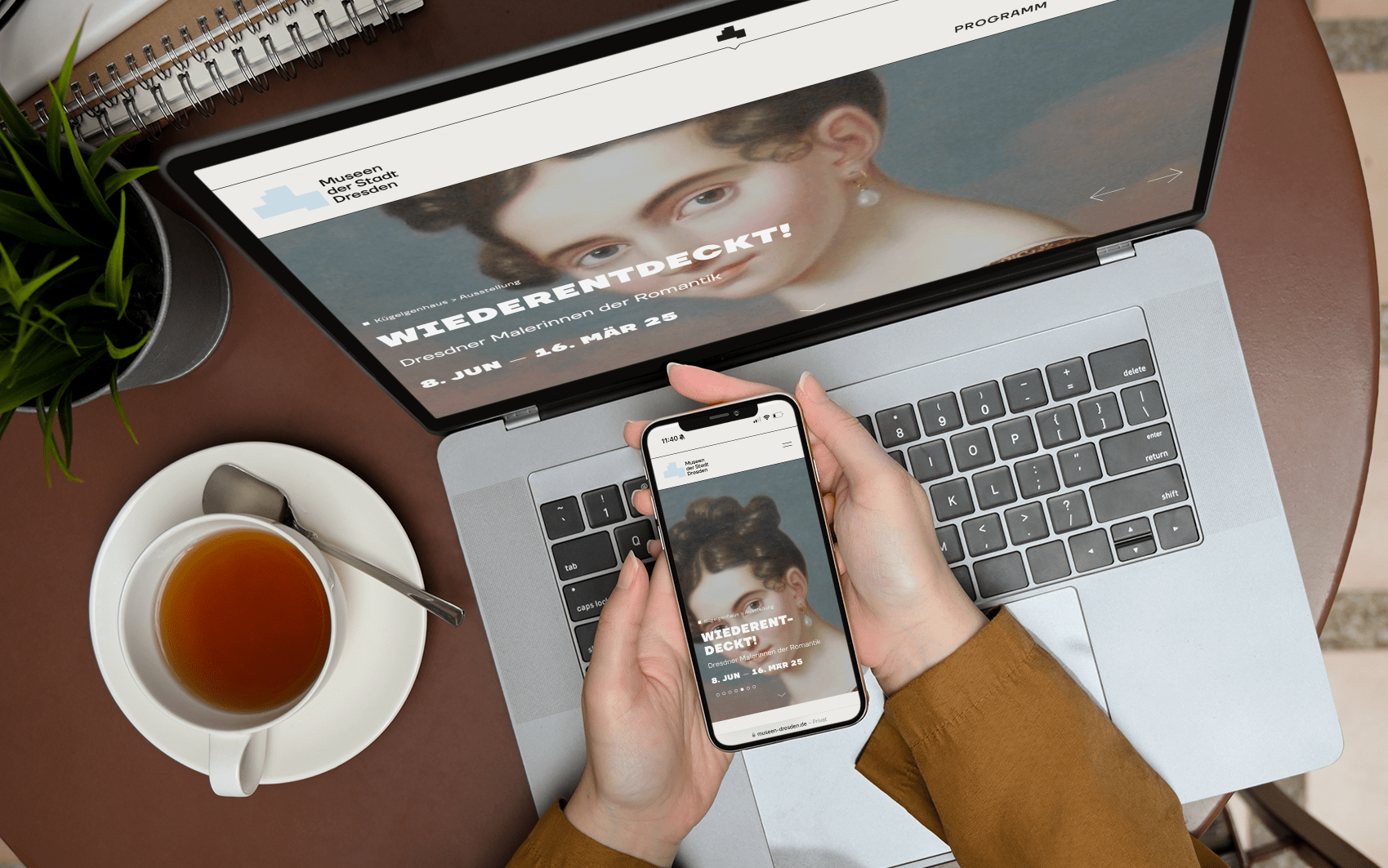 KI generiert: Das Bild zeigt eine Person, die ein Smartphone in der Hand hält, während ein Laptop mit einer Website des Museums der bildenden Künste Leipzig geöffnet ist, auf der eine Ausstellung namens "WIEDERENTDECKT! Dresdner Malerinnen der Romantik" präsentiert wird. Auf einem Tisch neben dem Laptop steht eine Tasse Tee.