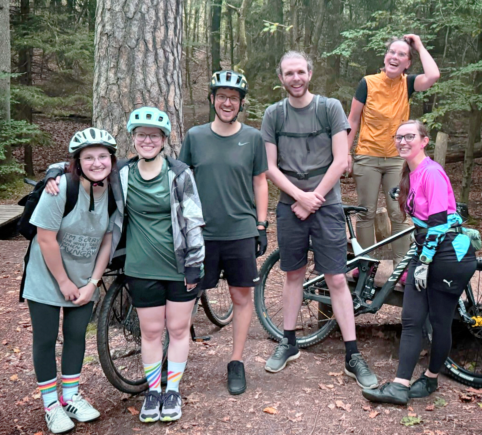 KI generiert: Eine Gruppe von sechs Personen in sportlicher Kleidung und Fahrradhelmen posiert mit Fahrrädern im Wald.