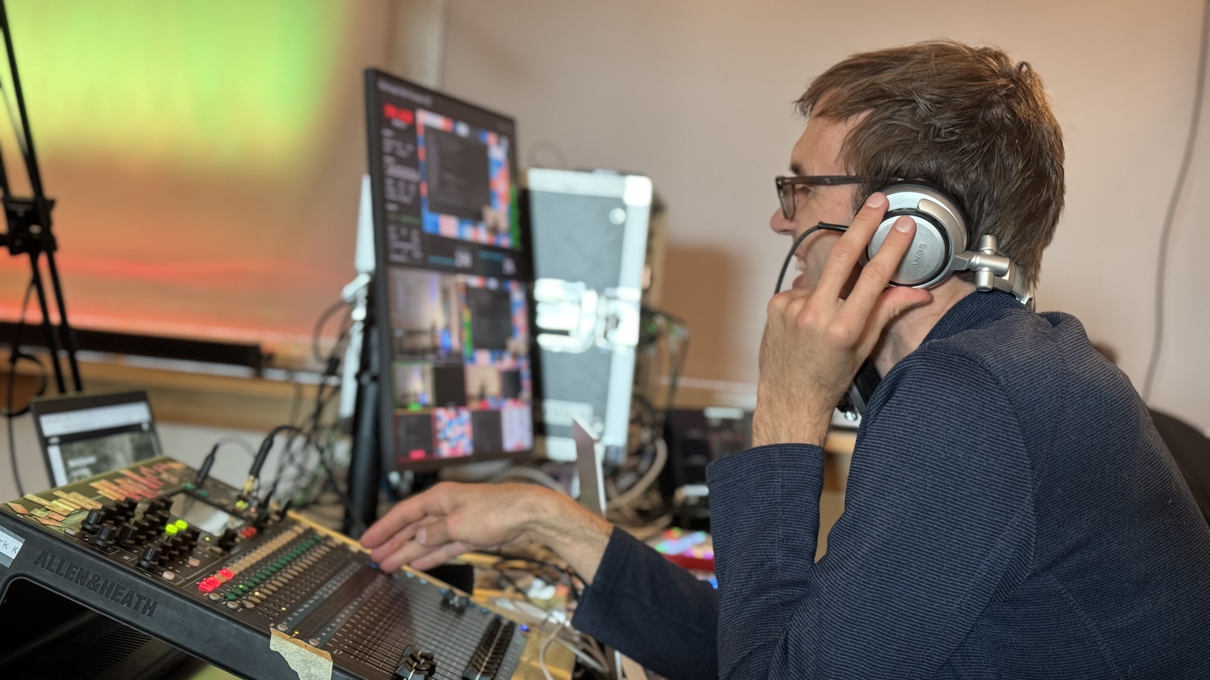 KI generiert: Das Bild zeigt eine Person, die an einem Mischpult in einem Studio arbeitet. Im Hintergrund sind Bildschirme mit visuellen Inhalten zu sehen.