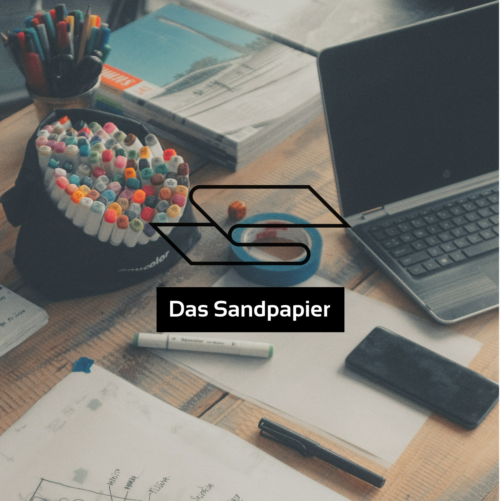 Ein Schreibtisch mit Laptop, Papier und bunten Stiften unter dem Podcast Logo "Das Sandpapier"