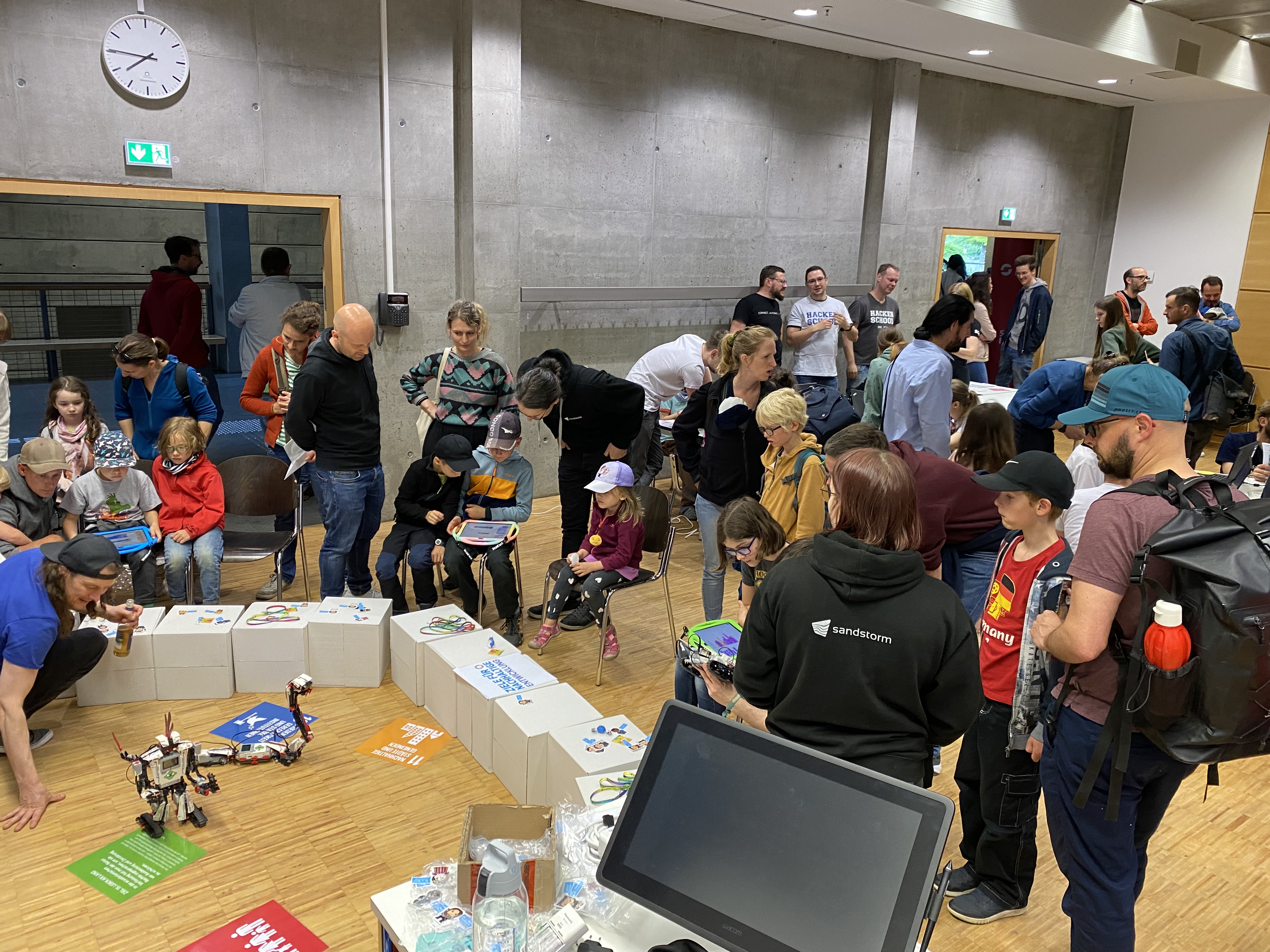 KI generiert: Das Bild zeigt eine Gruppe von Menschen, darunter viele Kinder, die in einem Raum zusammengekommen sind, möglicherweise zu einer Veranstaltung oder einem Workshop. Auf dem Boden sind Roboter und diverse Karten zu sehen, die von den Anwesenden betrachtet werden.