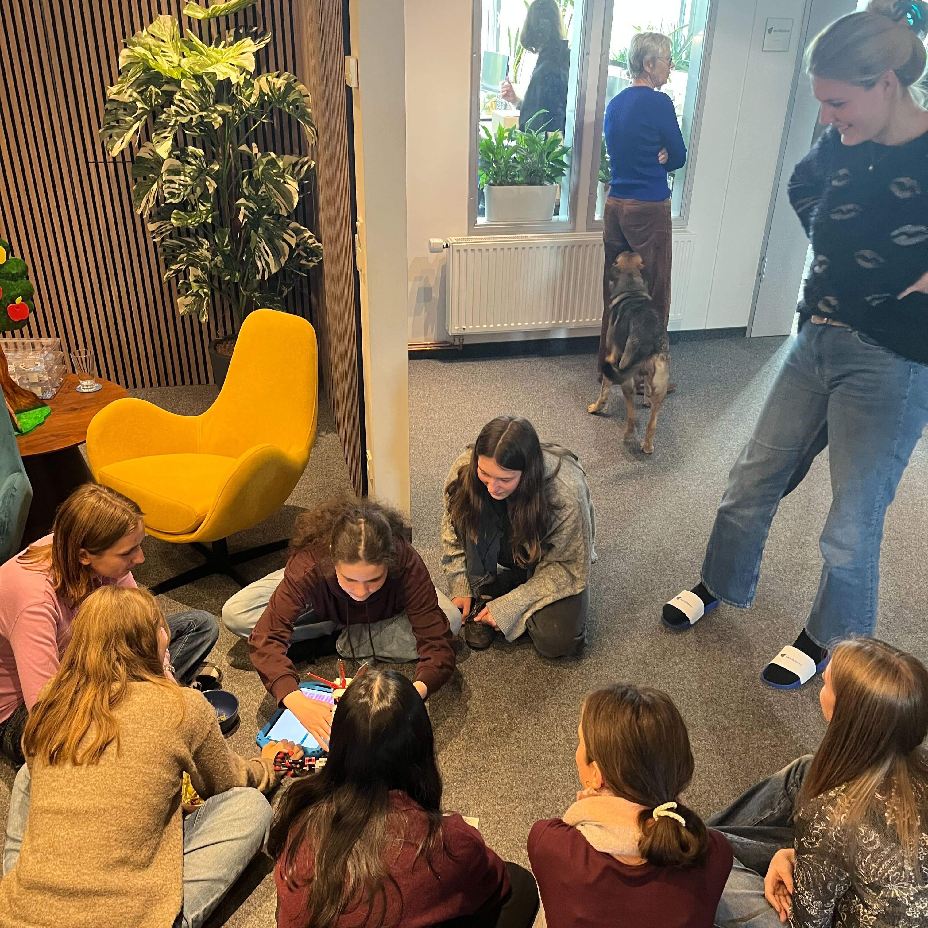 KI generiert: Eine Gruppe von Menschen sitzt auf dem Boden in einem Raum und konzentriert sich auf ein Tablet, während im Hintergrund jemand mit einem Hund steht.