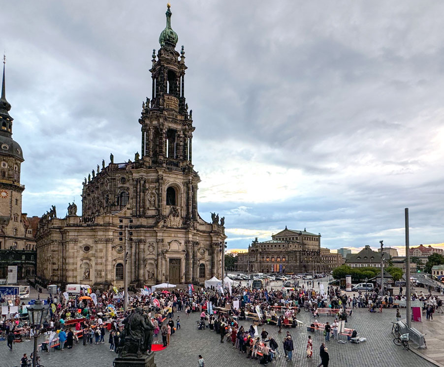 KI generiert: Das Bild zeigt einen belebten öffentlichen Platz in einer Stadt mit einer beeindruckenden historischen Kirche im Hintergrund. Zahlreiche Menschen tummeln sich auf dem Platz, möglicherweise auf einem Markt oder Festival.