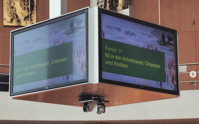 KI generiert: Das Bild zeigt einen großen Bildschirm, auf dem ein Forum zum Thema "KI in der Arbeitswelt: Chancen und Risiken" angekündigt wird. Der Bildschirm befindet sich in einem modernen Innenraum.