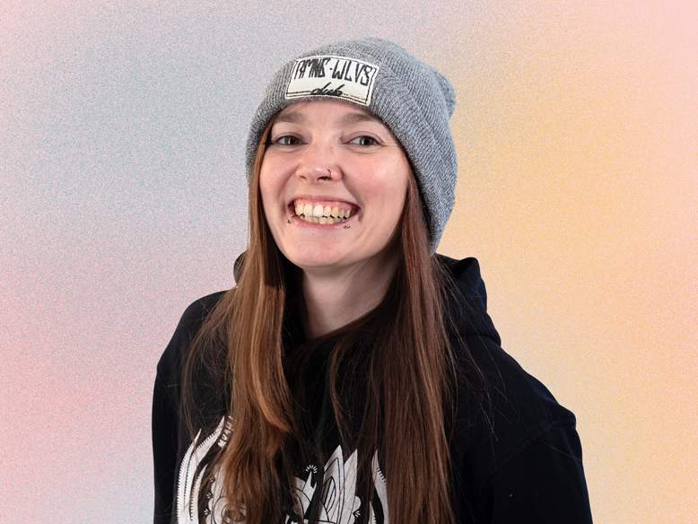 KI generiert: Das Bild zeigt eine lächelnde Person mit langem, braunem Haar, die einen grauen Beanie mit einer Aufschrift trägt und eine schwarze Jacke anhat.