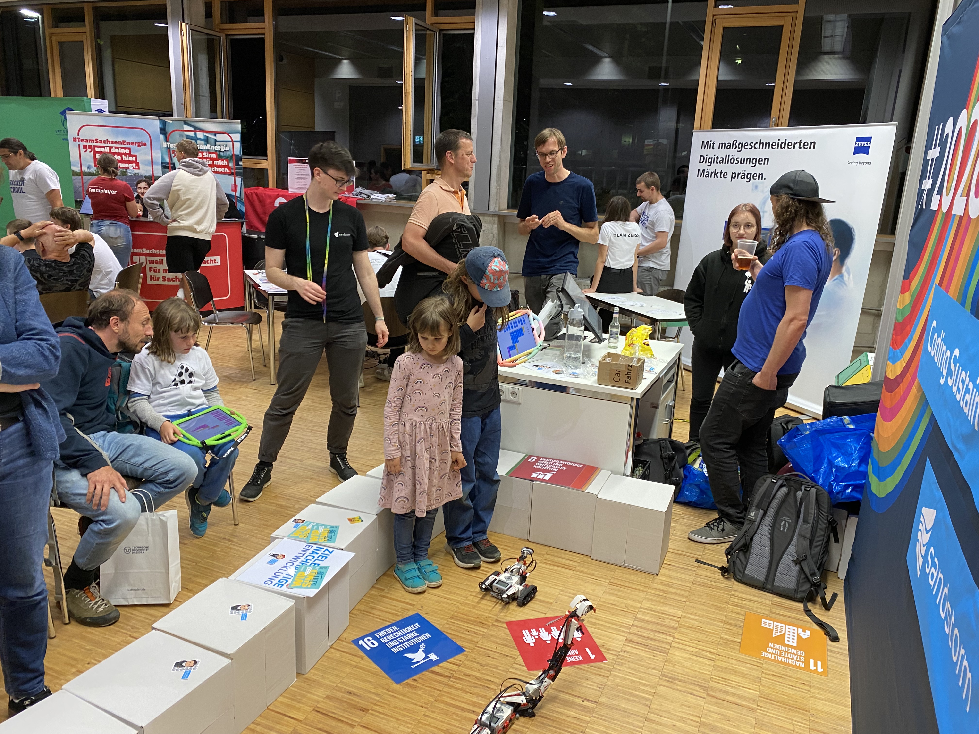 KI generiert: Auf dem Bild befindet sich eine Gruppe von Menschen in einem Raum, die offenbar an einem technischen oder digitalen Workshop teilnehmen. Kinder und Erwachsene sind um Roboter und Plakate versammelt, auf denen Nachhaltigkeitsziele abgebildet sind.