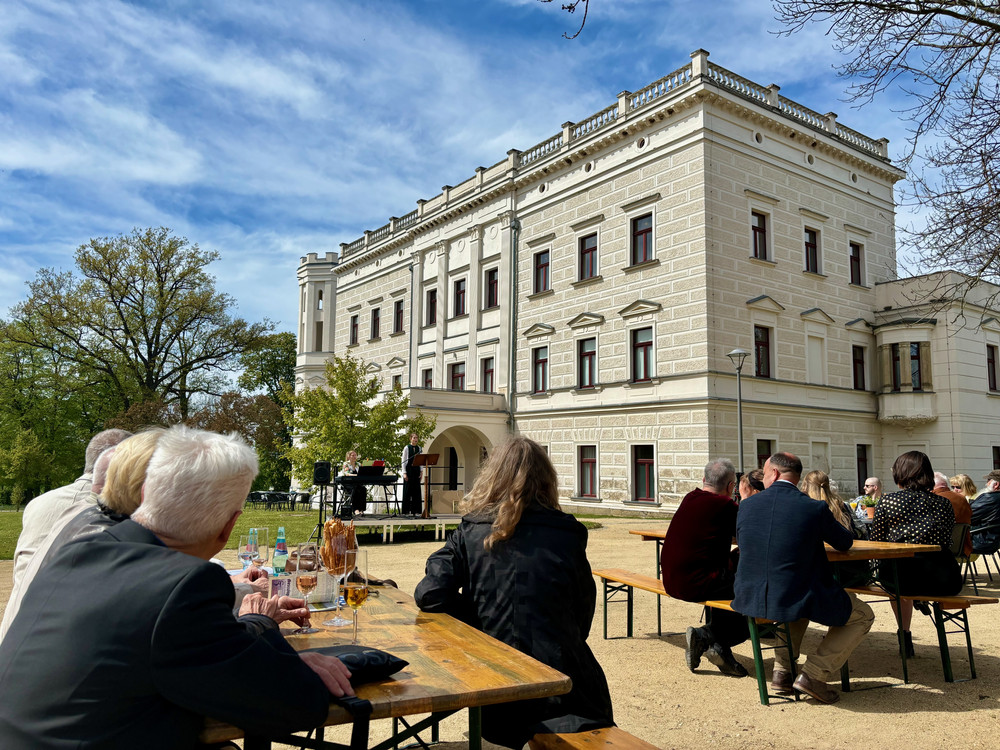 KI generiert: Das Bild zeigt eine Gruppe von Menschen, die vor einem historischen Gebäude im Freien an Biertischen sitzen. Im Hintergrund tritt eine Musikgruppe auf einer kleinen Bühne auf.
