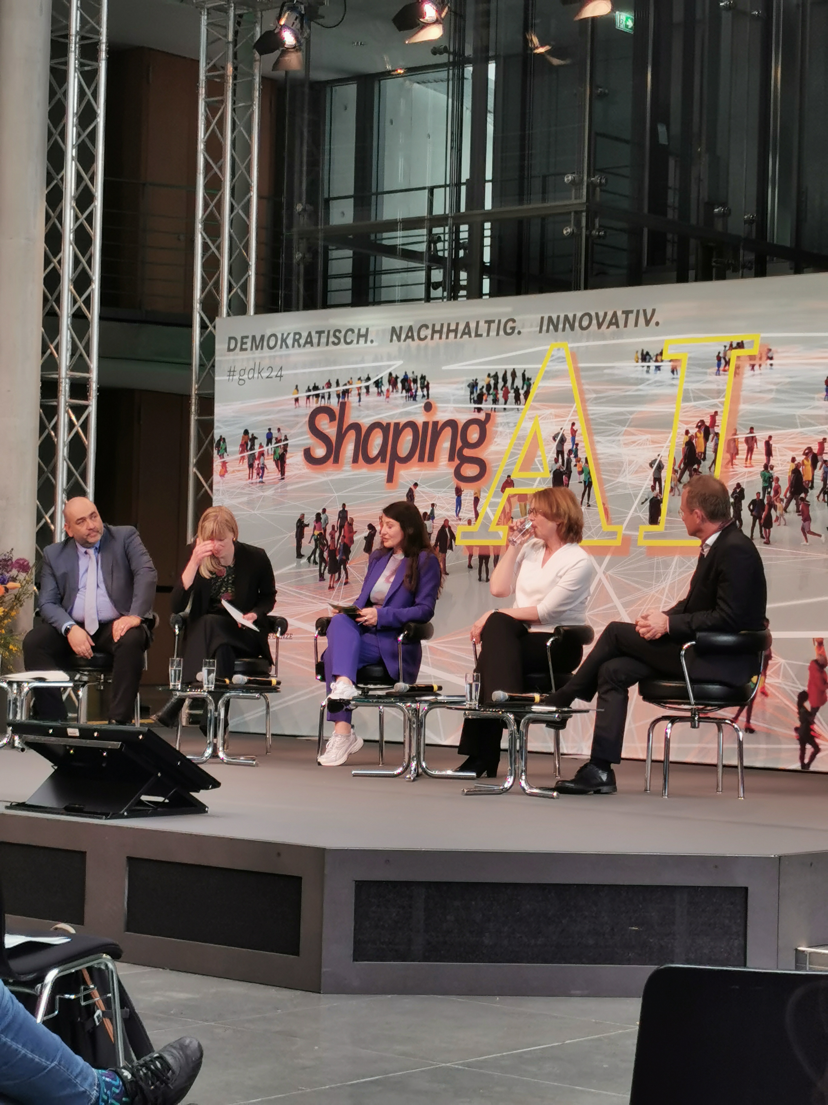 KI generiert: Das Bild zeigt eine Podiumsdiskussion mit fünf Personen vor einem Hintergrund mit der Aufschrift "Shaping AI". Das Thema der Veranstaltung scheint sich um die Gestaltung und Zukunft von Künstlicher Intelligenz zu drehen.