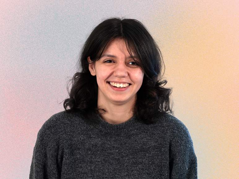 KI generiert: Das Bild zeigt eine lächelnde Person mit dunklen, welligen Haaren, die einen grauen Pullover trägt und vor einem Hintergrund mit sanftem Farbverlauf von Pink zu Orange steht.