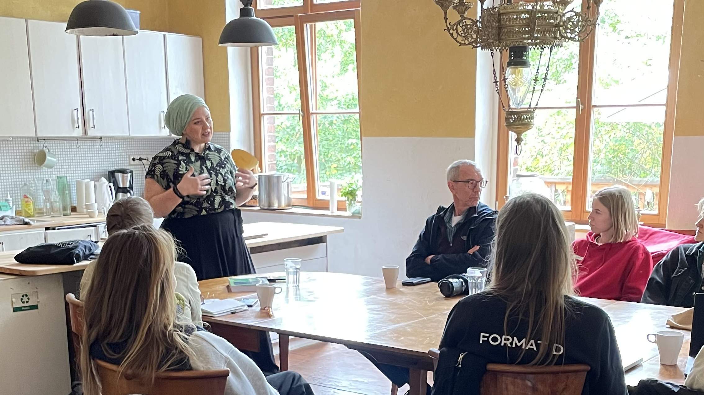 KI generiert: Eine Person spricht in einer gemütlichen Küchenszene zu einer Gruppe sitzender Menschen.