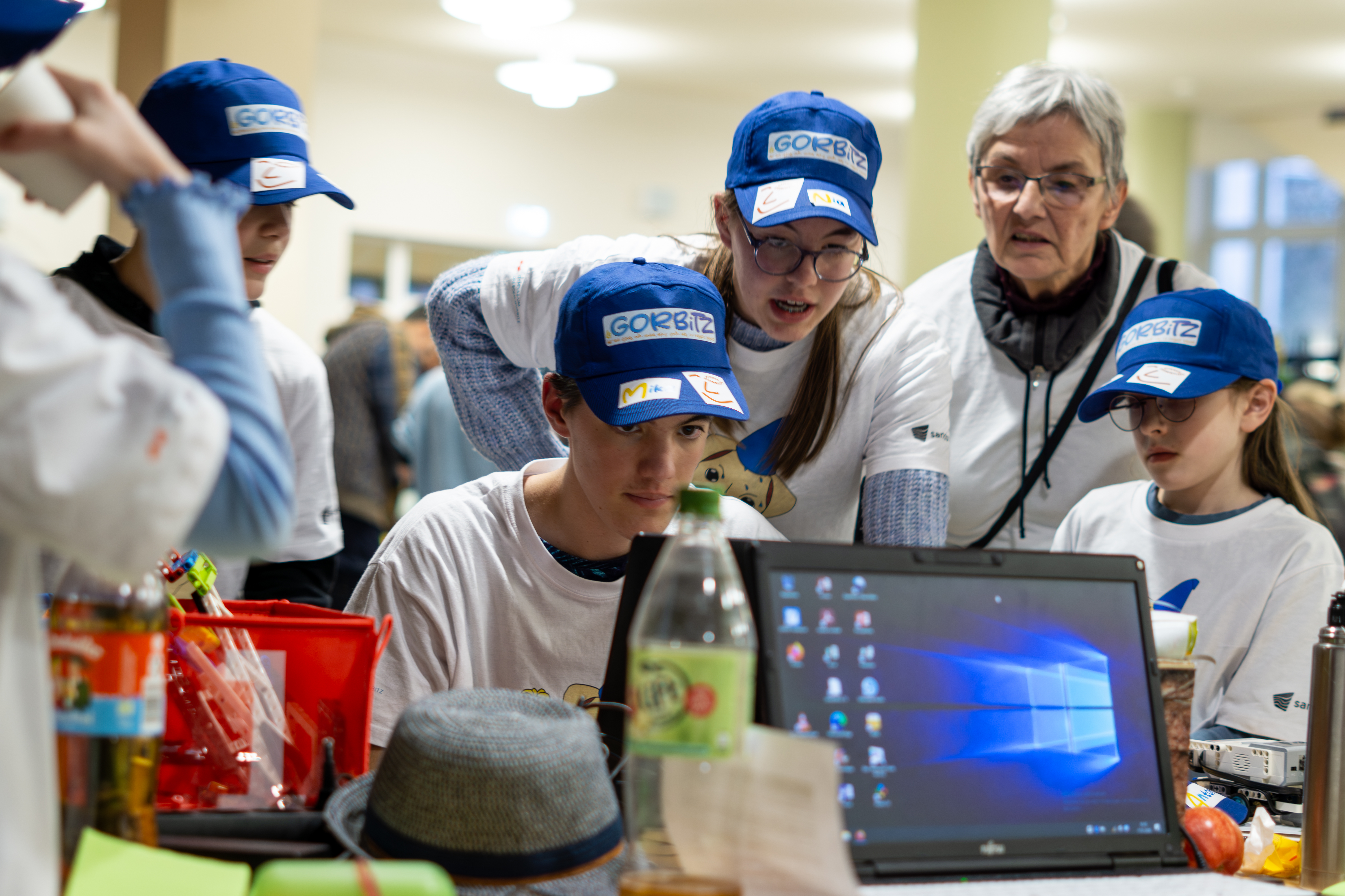 KI generiert: Das Bild zeigt eine Gruppe von Menschen, die sich um einen Laptop versammelt haben, wobei einige von ihnen blaue Kappen mit der Aufschrift "GORBIZ" tragen. Sie scheinen in ein gemeinsames Projekt oder eine Diskussion vertieft zu sein, möglicherweise in einem Bildungs- oder Gemeinschaftszentrum.