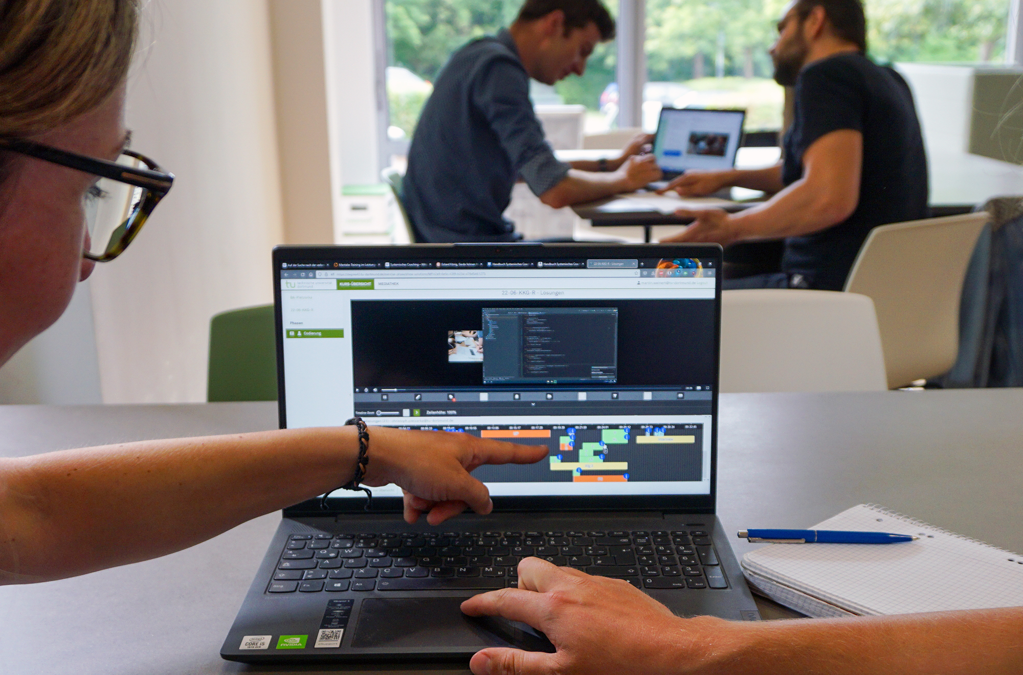 KI generiert: Das Bild zeigt eine Person, die auf den Bildschirm eines Laptops deutet, während im Hintergrund zwei Personen an einem Tisch sitzen und ebenfalls mit einem Laptop arbeiten. Der Bildschirm zeigt eine Software mit einer Zeitleiste, möglicherweise für Video- oder Audiobearbeitung.