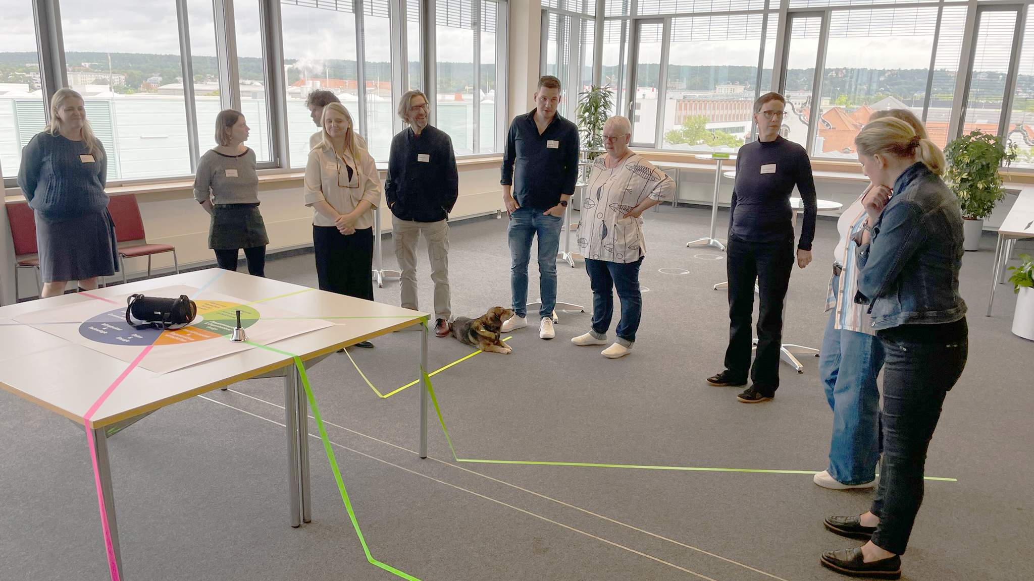 KI generiert: Eine Gruppe von Menschen steht in einem Kreis um einen Tisch in einem Büro, auf dem bunte Linien verlaufen.