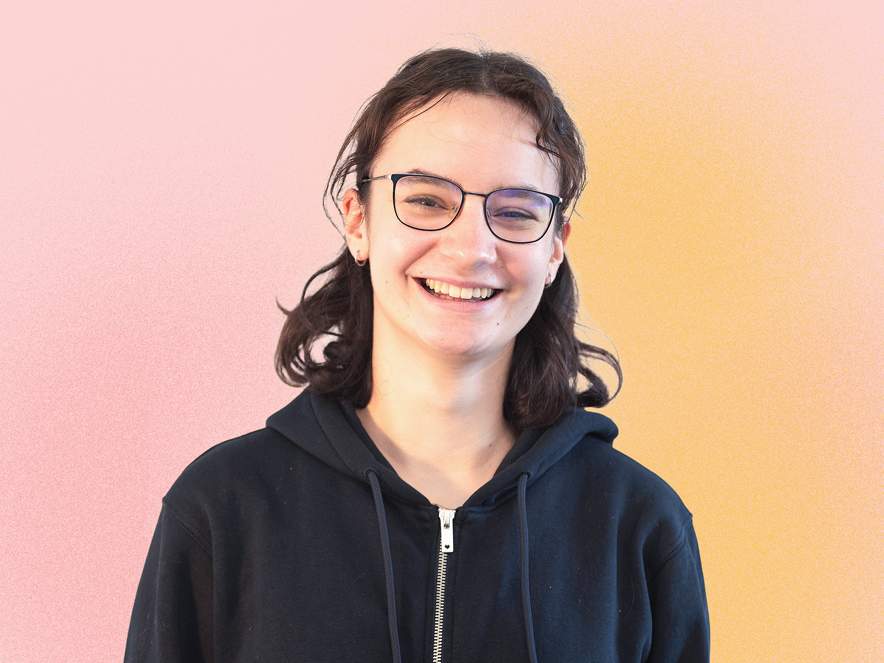 KI generiert: Das Bild zeigt eine lächelnde Person mit Brille und dunklen Locken, die einen schwarzen Kapuzenpulli trägt, vor einem pastellfarbenen Hintergrund.
