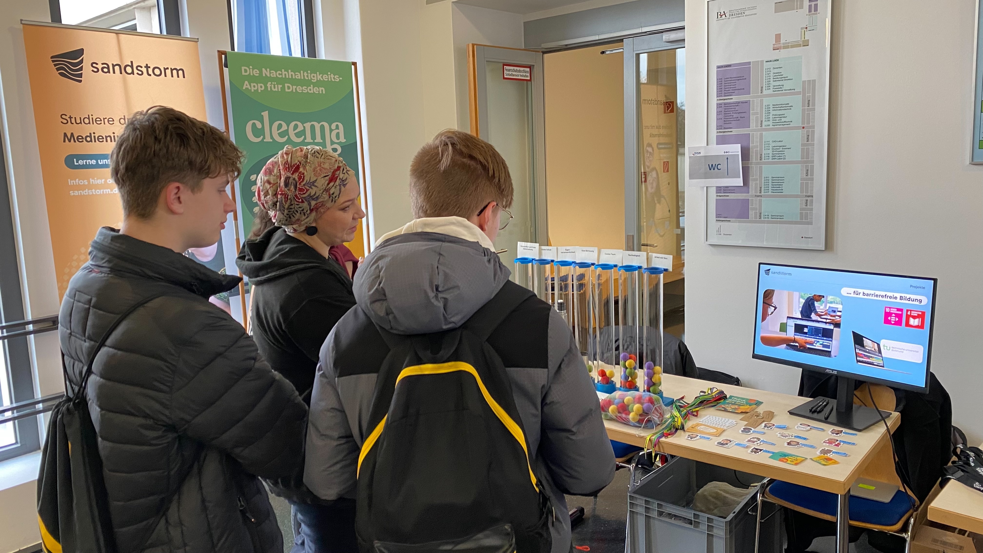 Karoline von Sandstorm und zwei interessierte Schüler schauen gemeinsam auf den Stand und die Präsentation zum Unternehmen