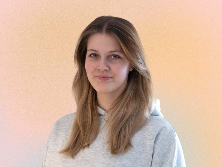 KI generiert: Das Bild zeigt eine lächelnde Frau mit langen, blonden Haaren, die vor einem hellen, einfarbigen Hintergrund steht.