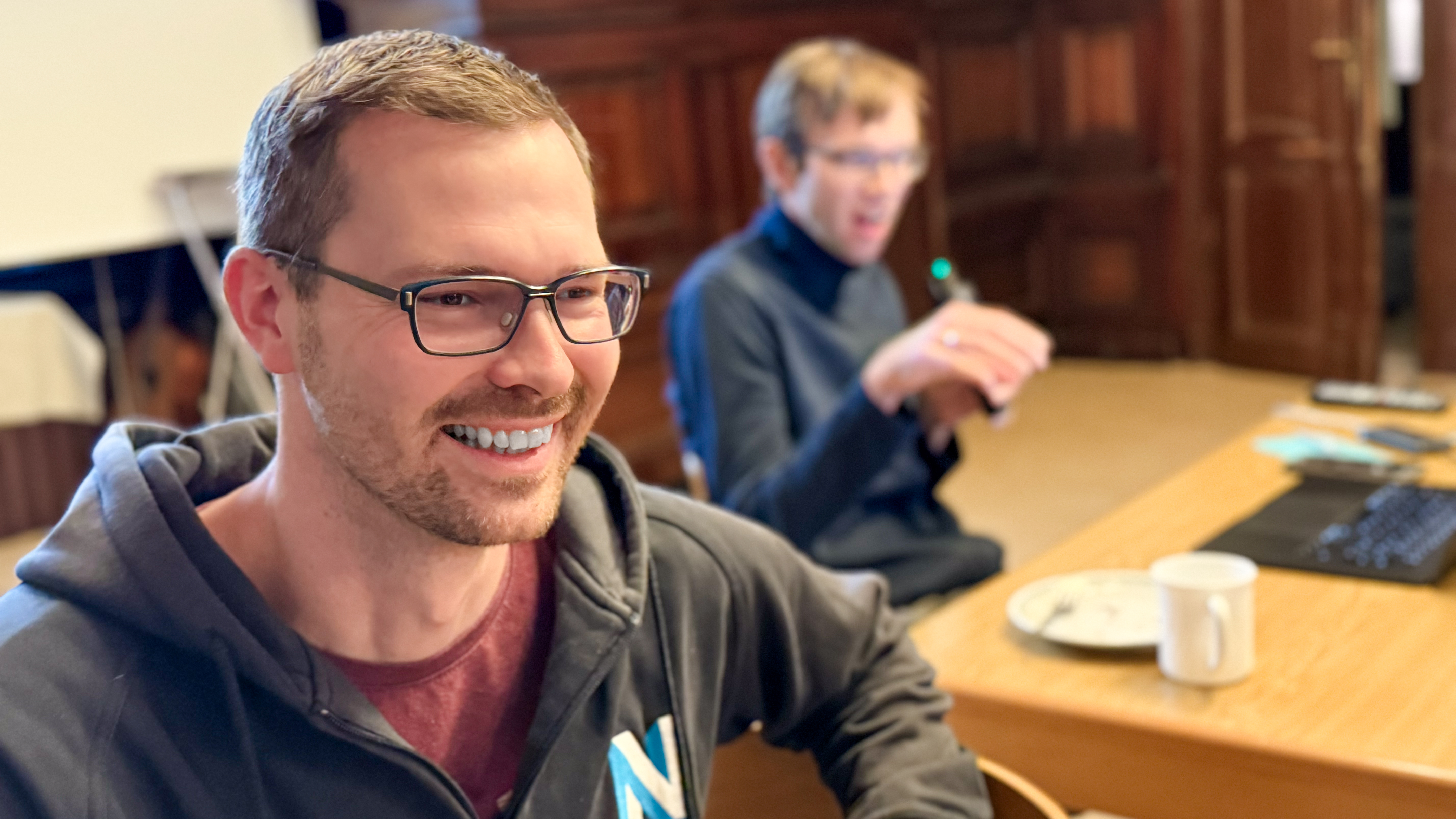 Das Bild zeigt Tobi und Sebastian beim Strategie-Workshop in Rathen, von denen einer im Vordergrund lächelt. Im Hintergrund sitzt eine Person an einem Tisch mit Laptop und Tasse.