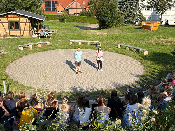 KI generiert: Das Bild zeigt eine Gruppe von Menschen, die in einem Freiluft-Amphitheater sitzt und zwei Personen in der Mitte der kreisförmigen Bühne beobachtet.