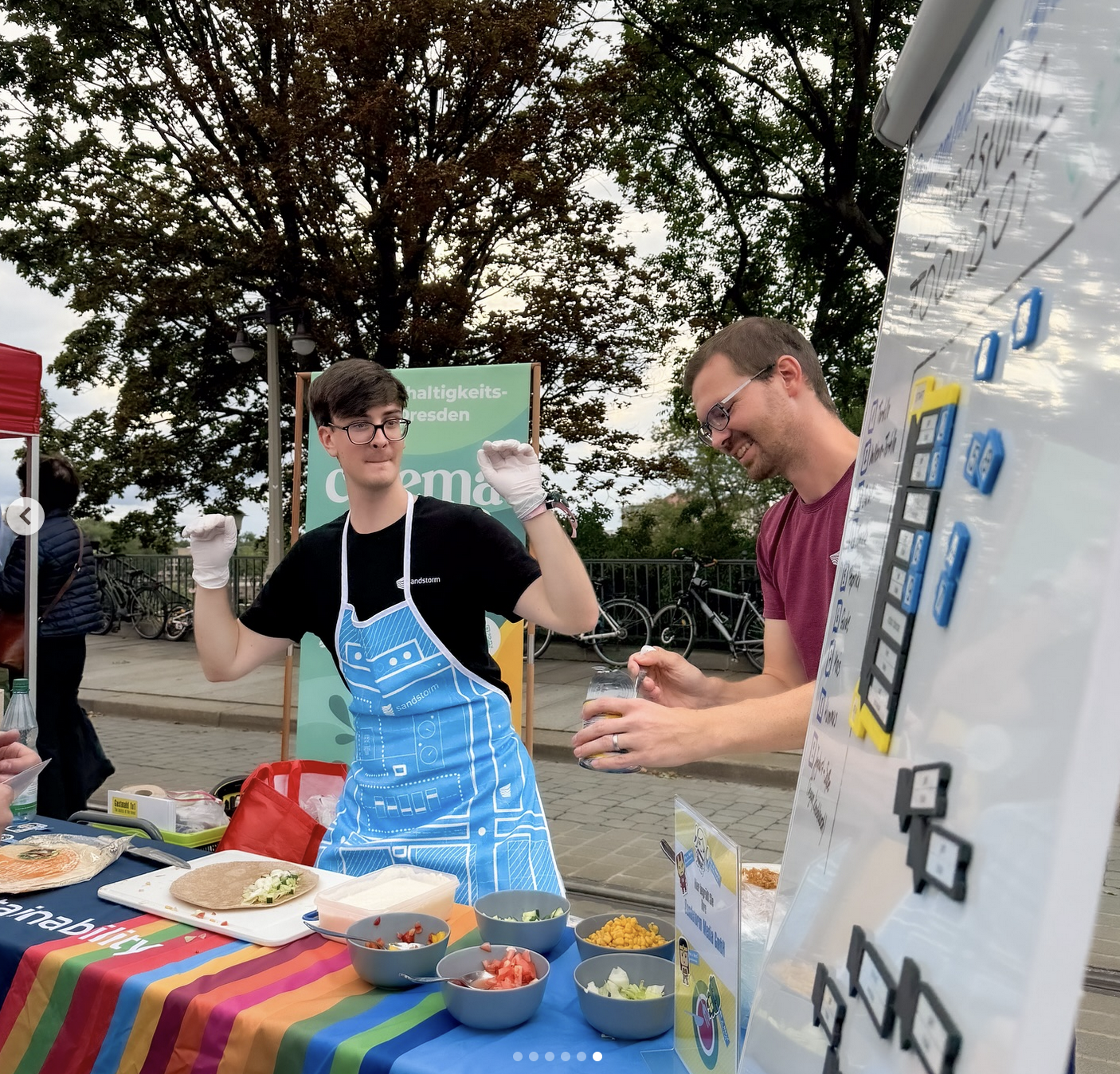 KI generiert: Das Bild zeigt zwei Personen an einem bunten Stand, die essbare Produkte zubereiten. Es scheint sich um eine Veranstaltung im Freien zu handeln, bei der Zutaten wie Gemüse angeboten werden.