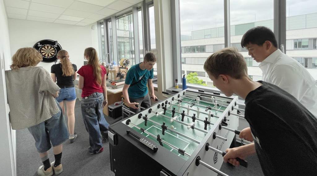 KI generiert: Der Hauptinhalt des Bildes zeigt eine Gruppe von Jugendlichen, die in einem hellen Raum mit großen Fenstern Foosball (Kicker) spielen und sich unterhalten.