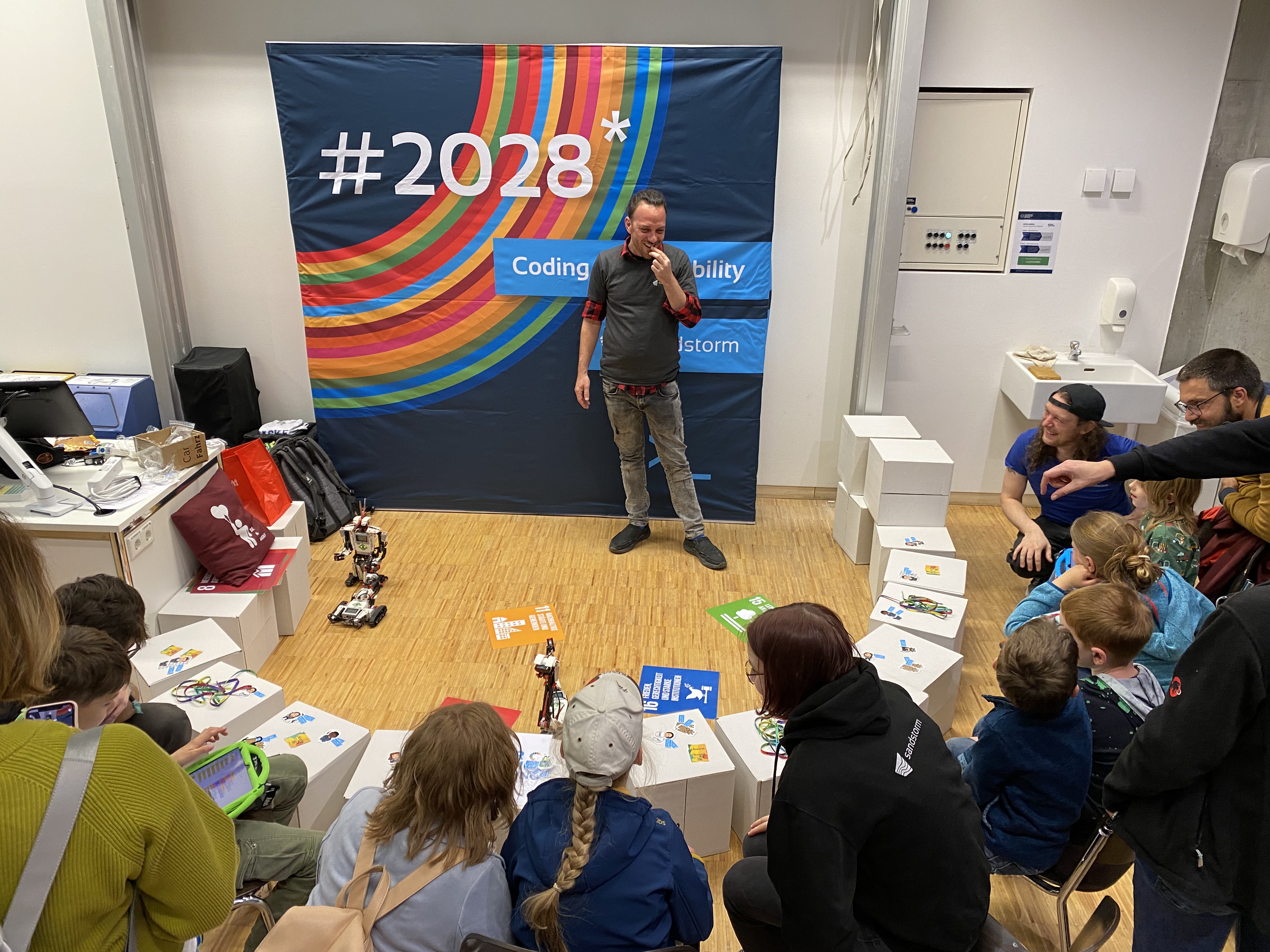 KI generiert: Ein Mann gibt eine Präsentation über Robotik vor einer Gruppe von Kindern in einem Raum mit einem bunten Banner im Hintergrund. Die Kinder sitzen im Halbkreis und scheinen aufmerksam zuzuhören.