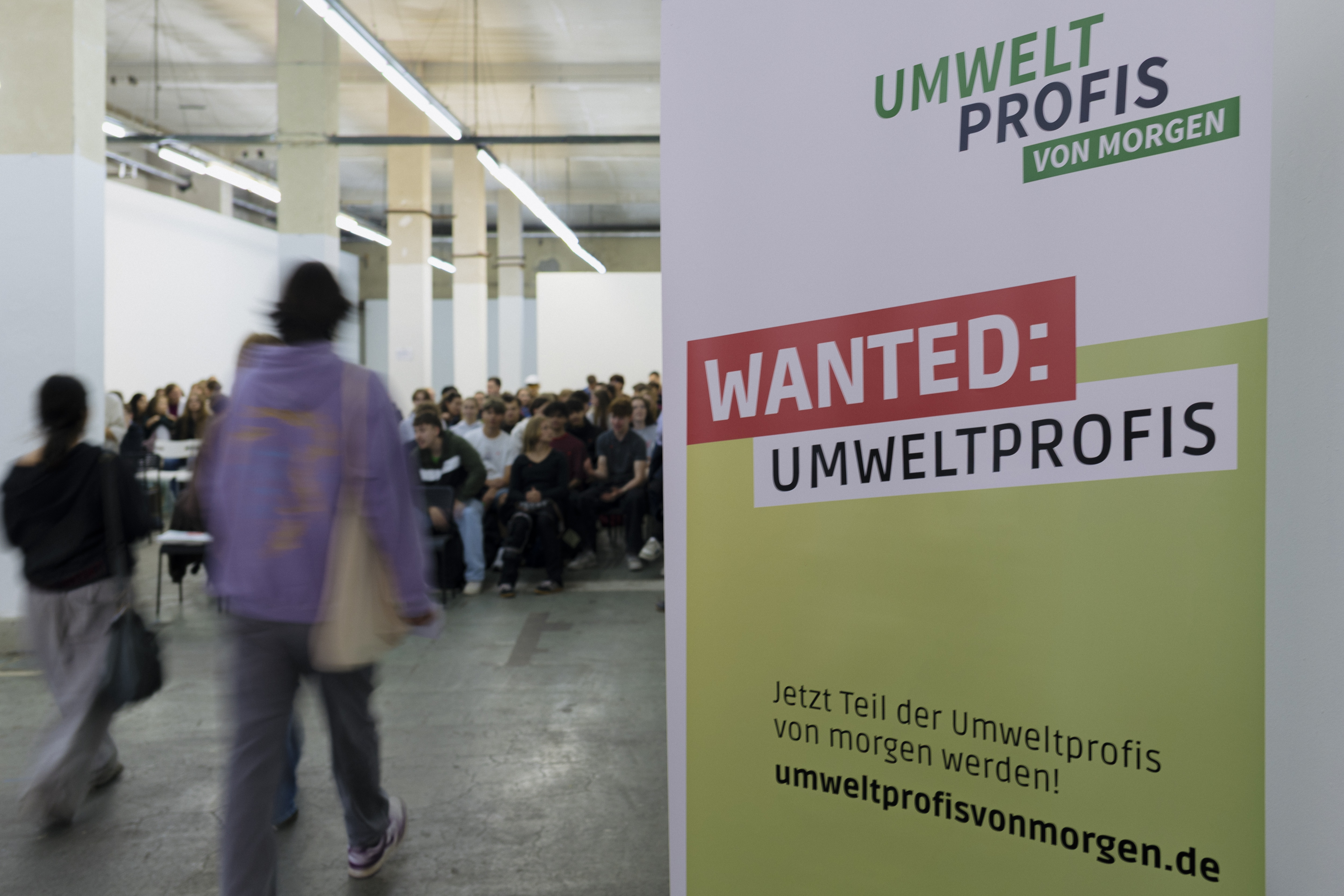 KI generiert: Das Bild zeigt eine Veranstaltung, bei der eine Gruppe von Menschen in einem Raum sitzt, während im Vordergrund ein Plakat mit der Aufschrift "WANTED: UMWELTPROFIS" und der Website "umweltprofisvonmorgen.de" zu sehen ist.