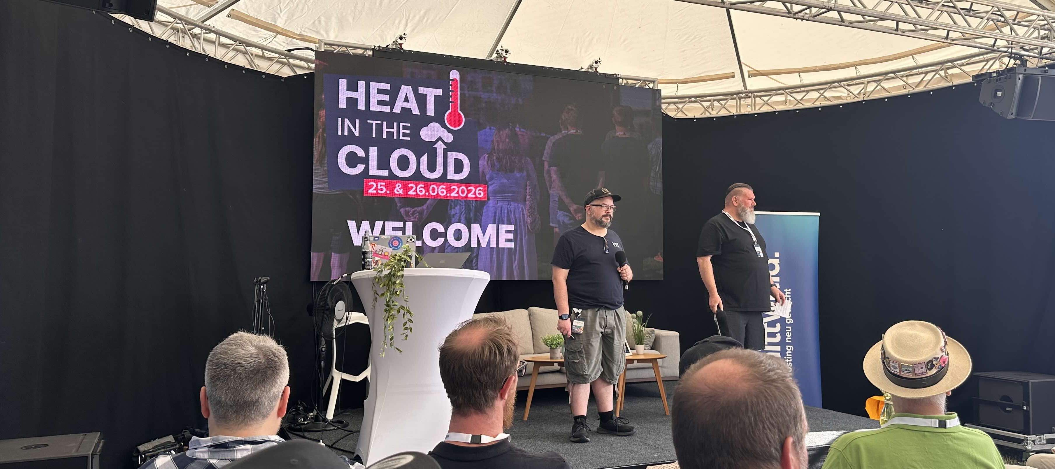 KI generiert: Das Bild zeigt zwei Männer auf einer Bühne, die vor einem Publikum sprechen, mit einem großen Bildschirm im Hintergrund, der die Begrüßung zu einer Veranstaltung namens "Heat in the Cloud" am 25. und 26. Juni 2026 anzeigt.