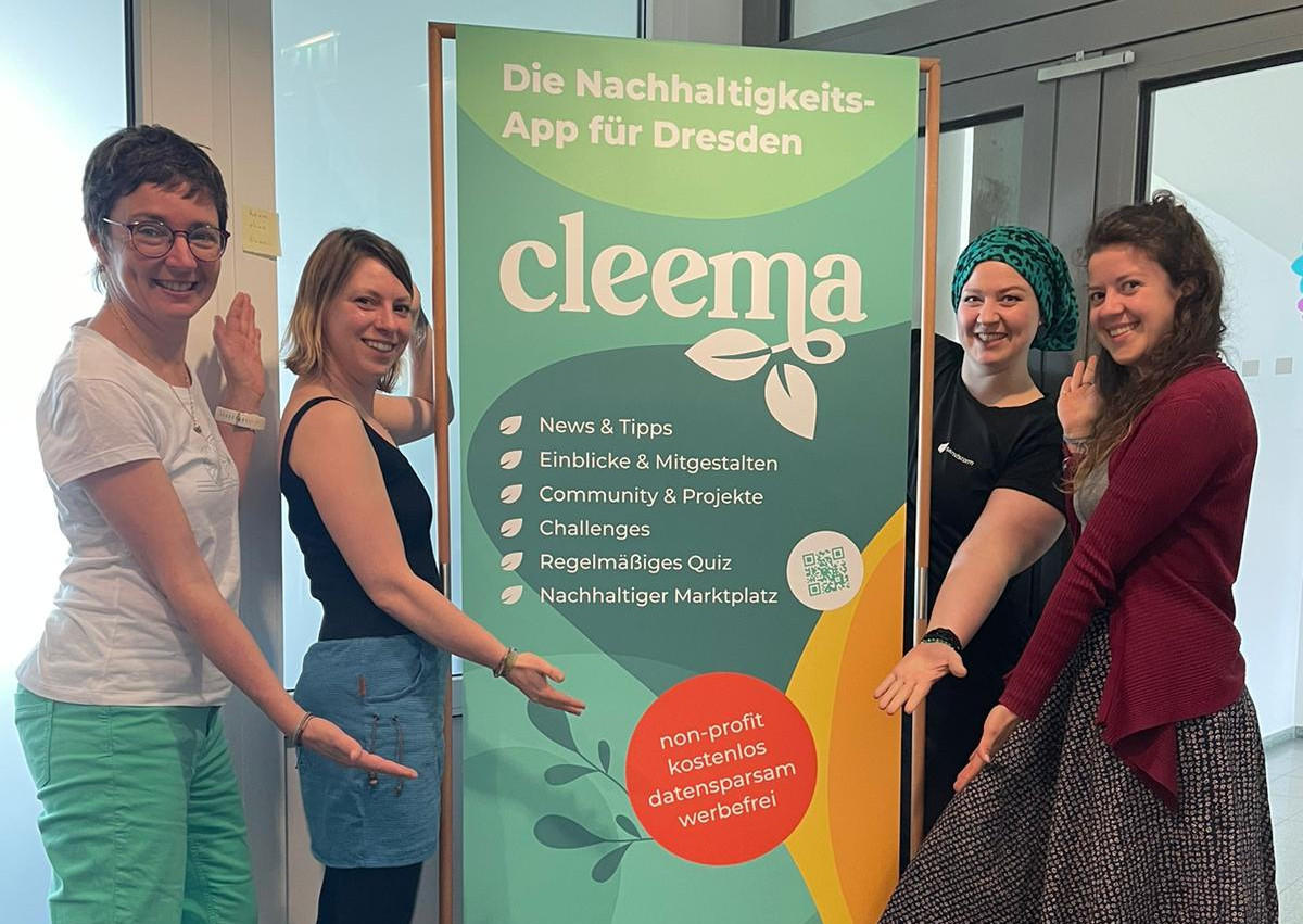 KI generiert: Das Bild zeigt vier Personen, die neben einem Werbebanner für die Nachhaltigkeits-App "cleema" in Dresden stehen. Sie lächeln in die Kamera und präsentieren das Banner.