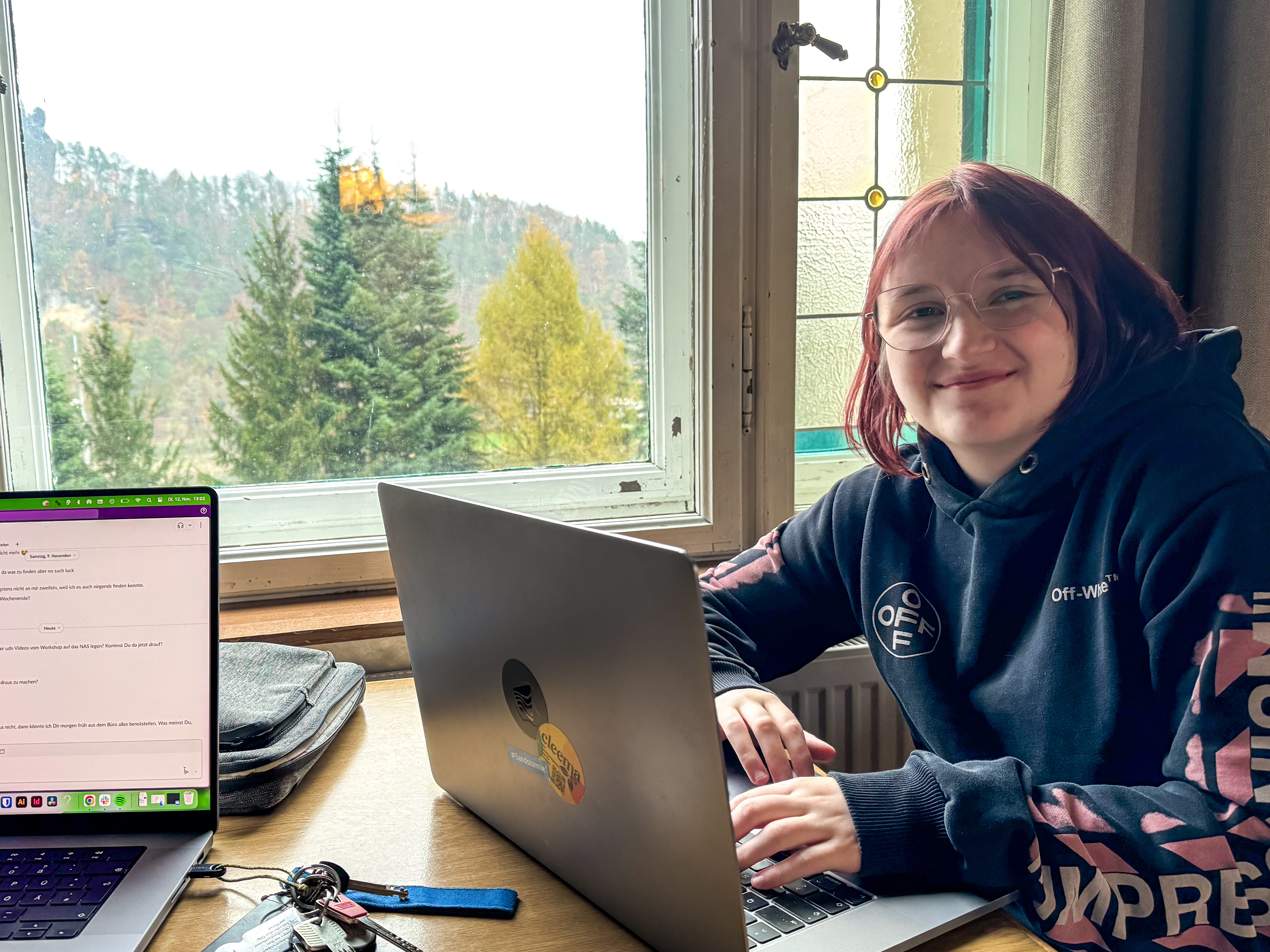 KI generiert: Eine Person sitzt an einem Tisch mit einem Laptop und lächelt, während sie aus einem Fenster auf eine bewaldete Landschaft blickt. Auf dem Tisch liegen außerdem ein zweiter Laptop und ein Schlüsselbund.