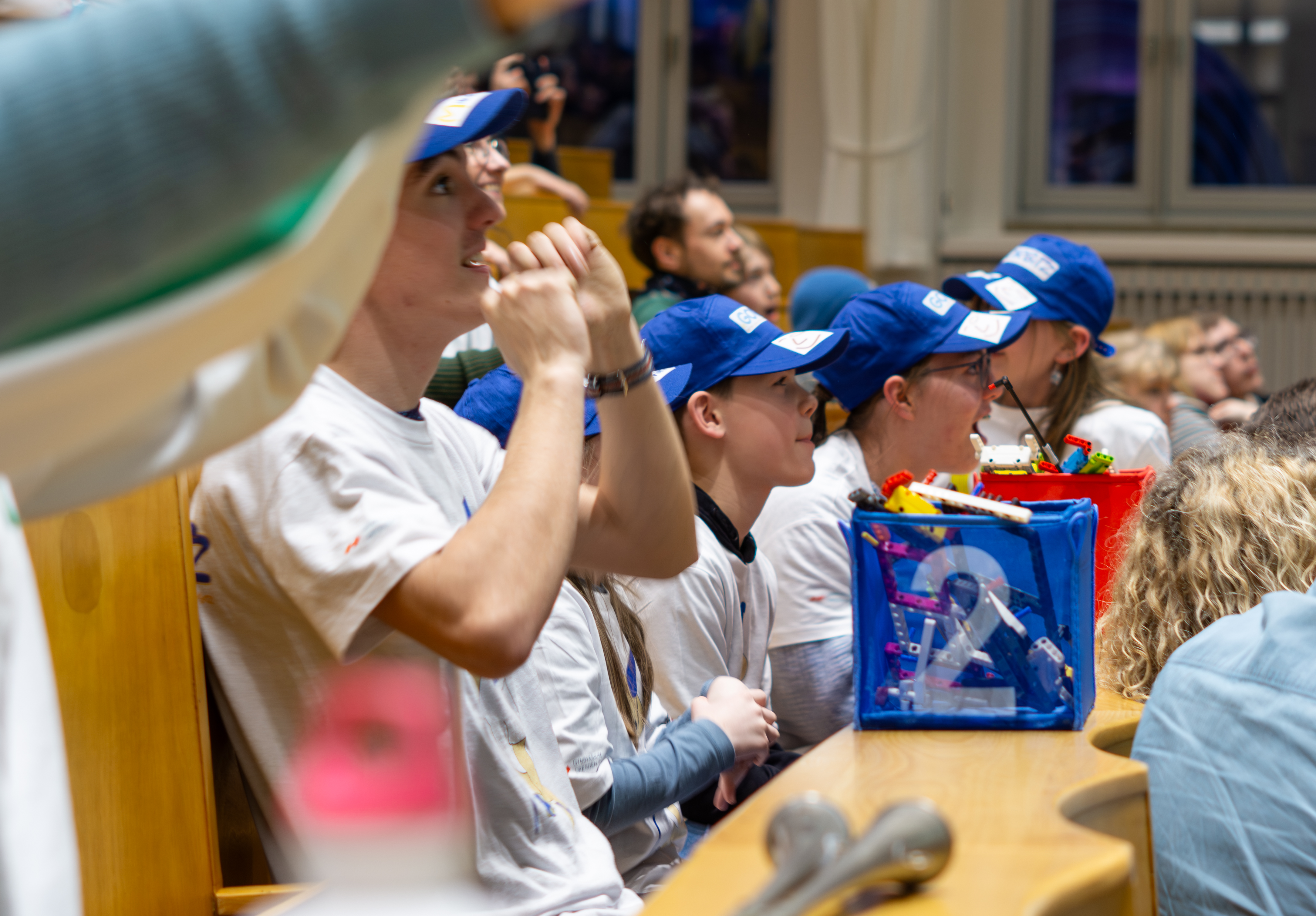 KI generiert: Das Bild zeigt eine Gruppe von Menschen, die in einem Raum mit Holzstühlen sitzen und aufmerksam einer Präsentation oder einem Vortrag folgen. Die meisten von ihnen tragen weiße T-Shirts und blaue Baseballkappen, was darauf hindeutet, dass sie möglicherweise Teil eines Teams oder einer Organisation sind. Einige von ihnen haben Notizbücher oder andere Materialien auf ihren Schreibtischen, was darauf hindeutet, dass sie sich auf eine aktive Teilnahme an der Veranstaltung vorbereitet haben. Der Raum scheint gut beleuchtet und hat große Fenster, die natürliches Licht hereinlassen.