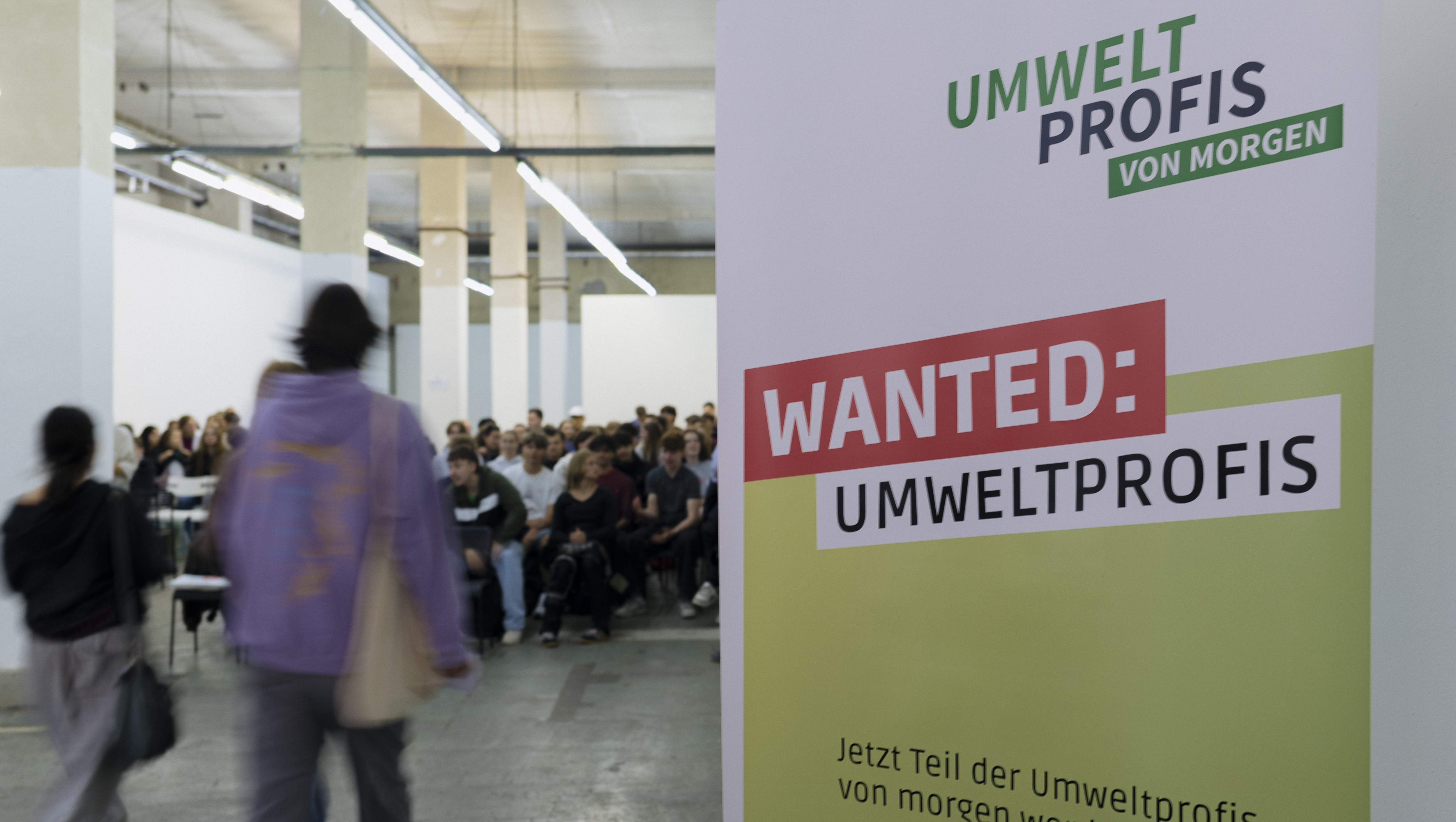 KI generiert: Das Bild zeigt eine Gruppe von Menschen in einem hellen, industriell gestalteten Raum, wobei im Vordergrund ein Plakat mit der Aufschrift "WANTED: UMWELTPROFIS" und "UMWELT PROFIS VON MORGEN" zu sehen ist.