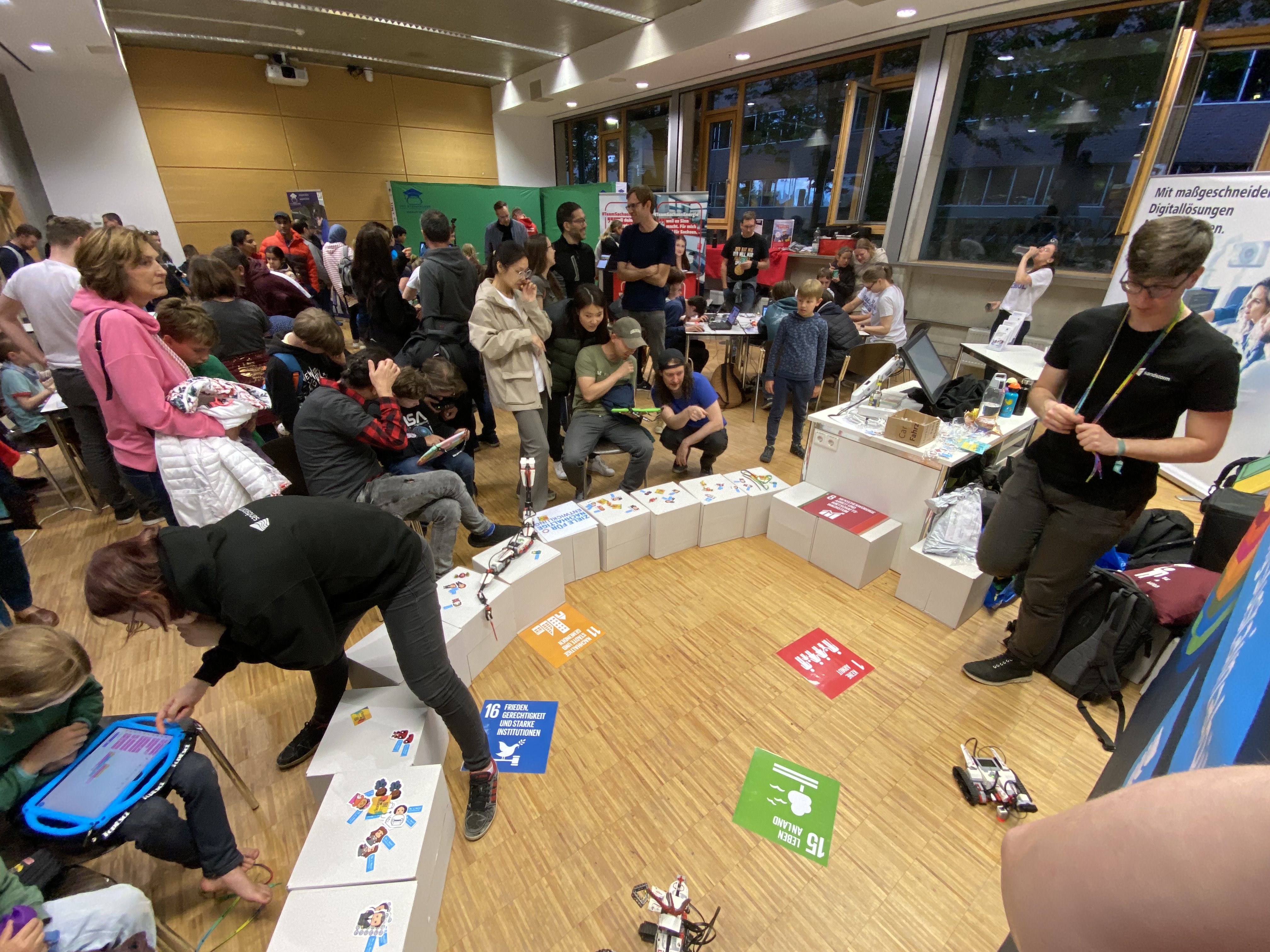 KI generiert: Das Bild zeigt eine Gruppe von Menschen, die sich um einen Bereich mit kleinen Robotern versammelt haben. Es herrscht reges Interesse und Aktivität, vermutlich im Rahmen eines Workshops oder einer Ausstellung zu Technologie oder Bildung.