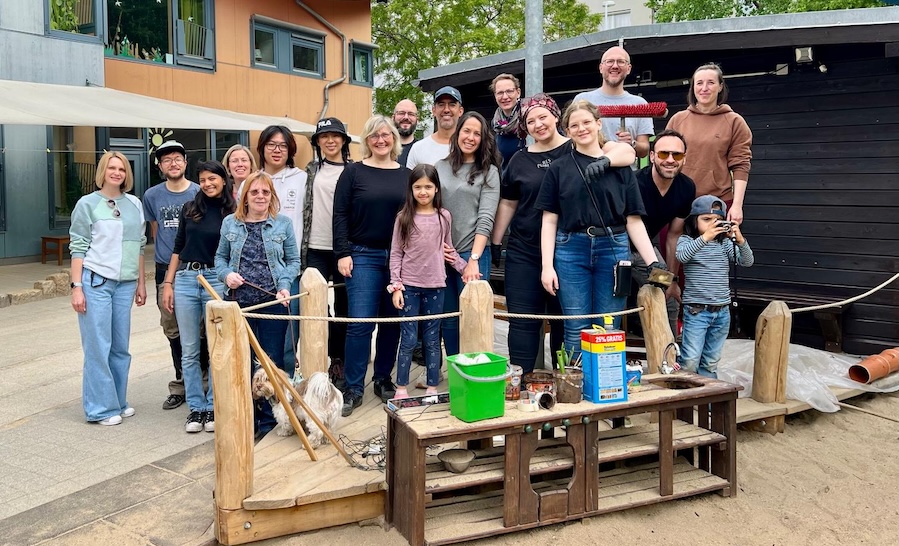 KI generiert: Eine Gruppe von Menschen steht zusammen draußen vor einem Gebäude, lächelnd und in ungezwungener Freizeitkleidung.