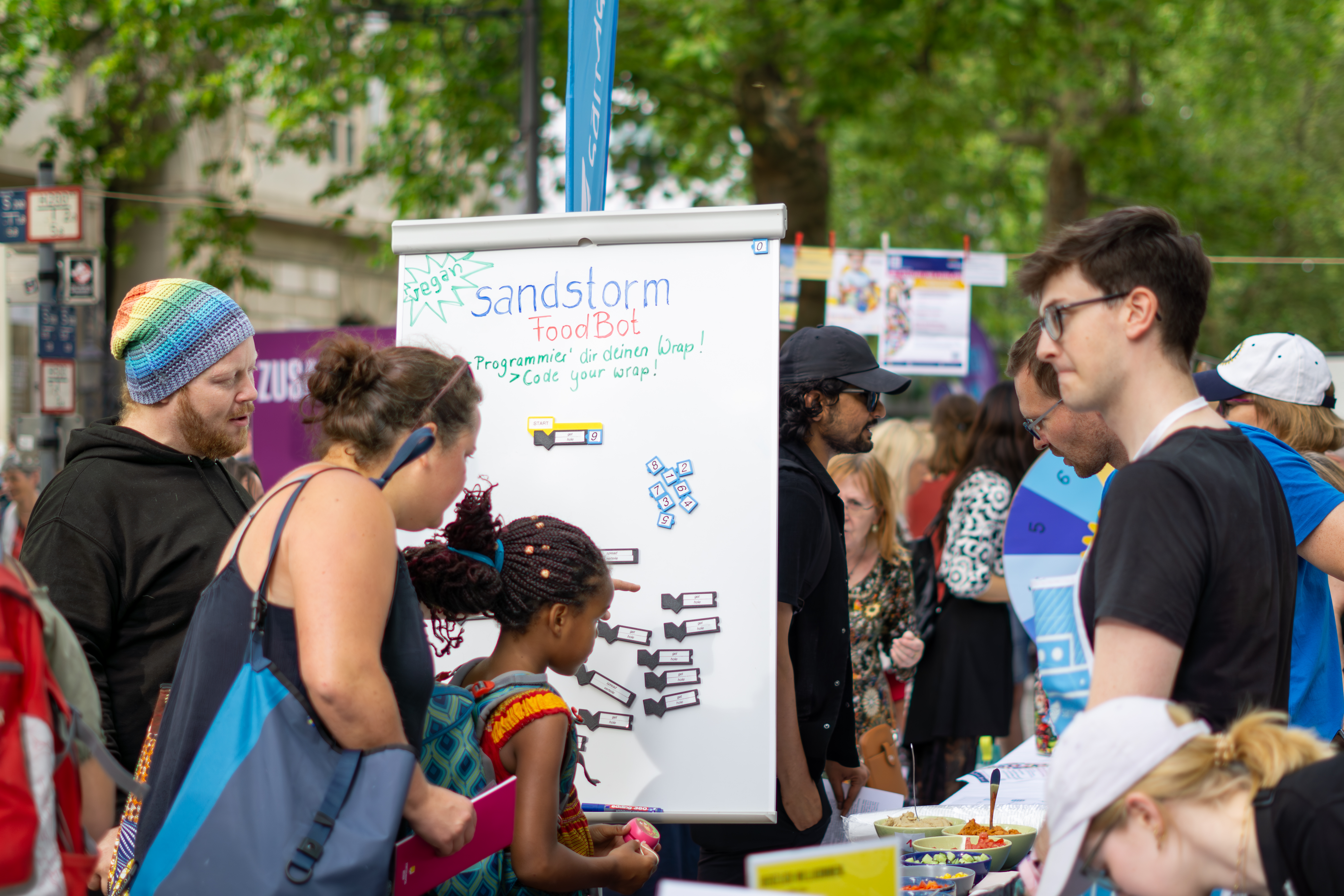 KI generiert: Menschen interagieren bei einer Veranstaltung oder einem Festival mit einem Stand, der mit "Sandstorm FoodBot" beschriftet ist.