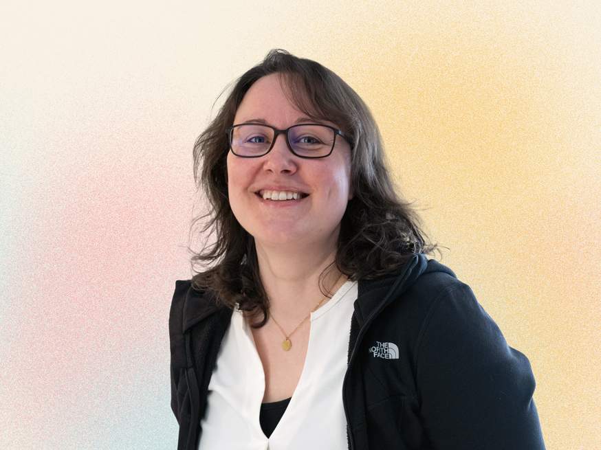 KI generiert: Das Bild zeigt eine lächelnde Frau mit Brille und lockigem Haar, die in die Kamera schaut. Sie trägt eine Jacke und ein helles Oberteil, während der Hintergrund unscharf ist.