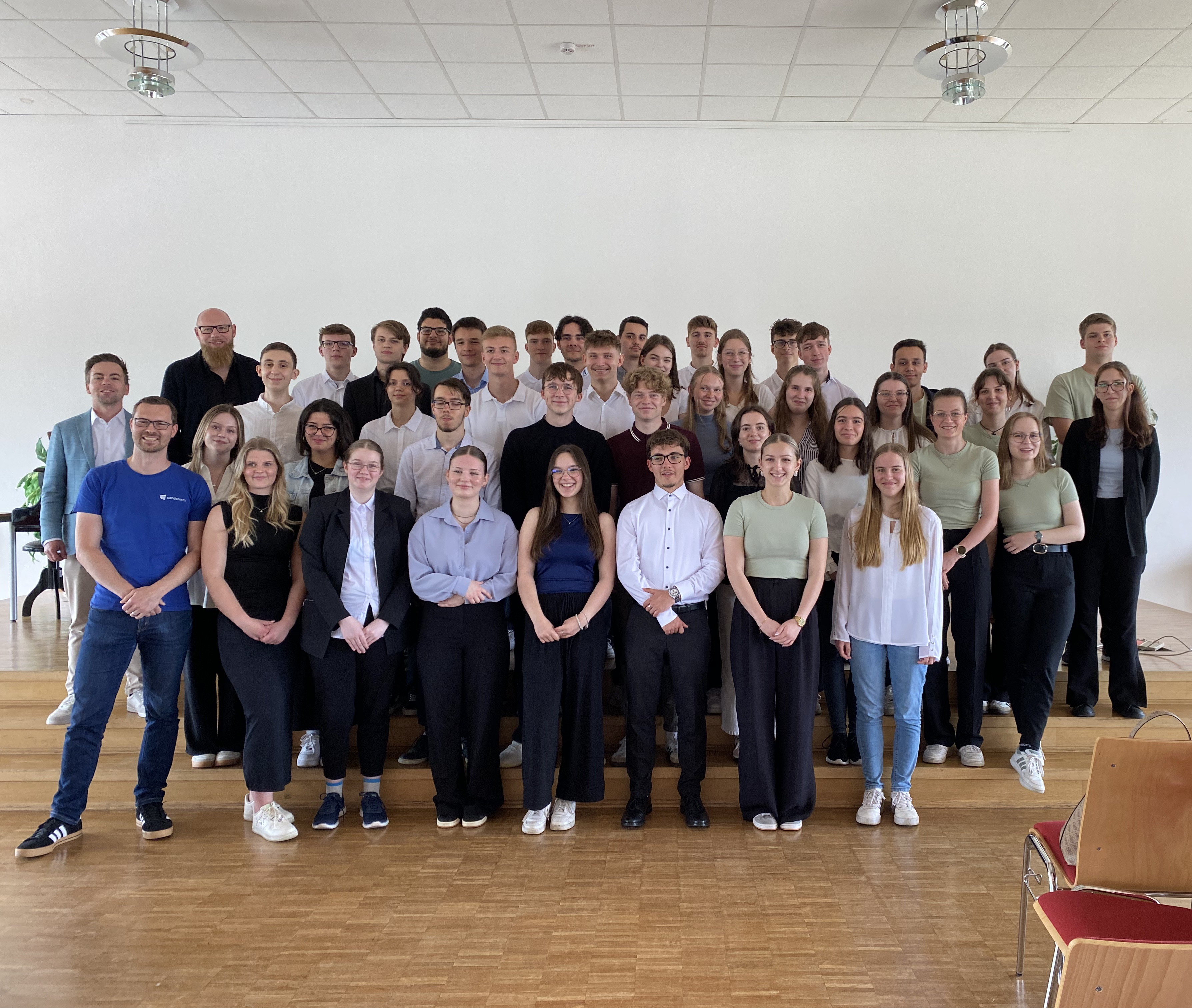 KI generiert: Eine große Gruppe von Menschen, meist jüngere Erwachsene, posiert für ein Gruppenfoto in einem hellen Raum mit Holzboden. Einige Personen stehen auf einer Art Bühne, was darauf hindeutet, dass es sich um eine formelle oder organisierte Veranstaltung handeln könnte.