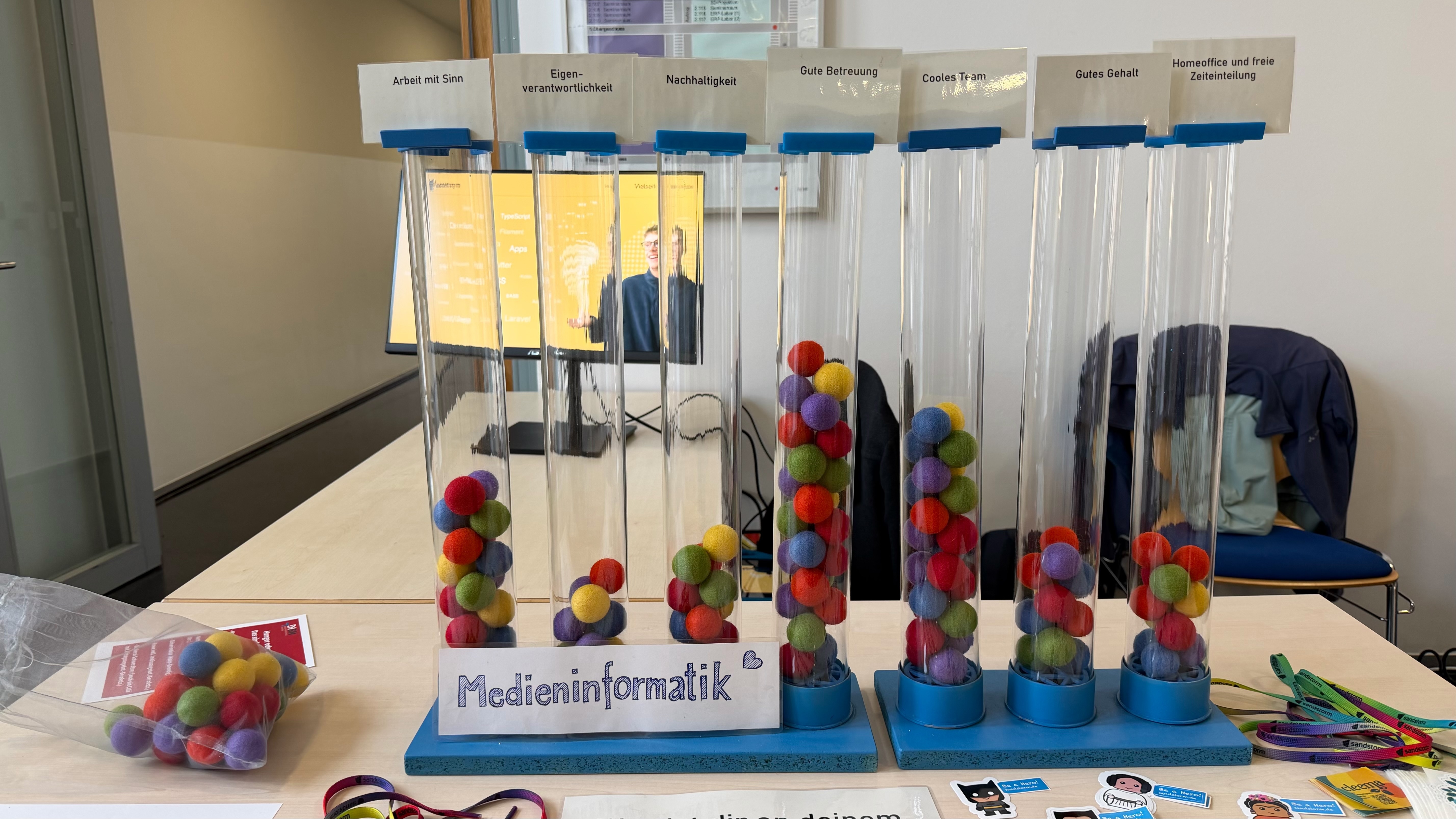 KI generiert: Das Bild zeigt einen Tisch mit mehreren durchsichtigen Röhren, die mit bunten Gummibärchen gefüllt sind, und einem Schild mit der Aufschrift "Medieninformatik".