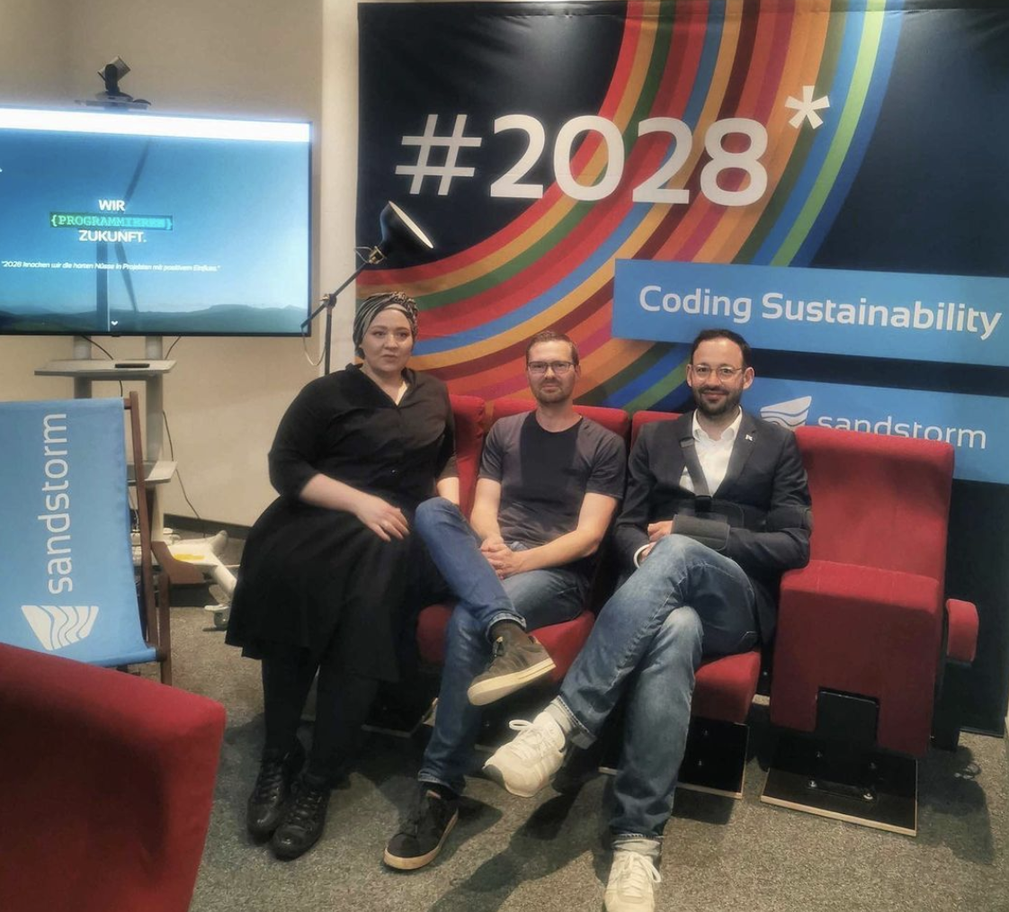 Karoline, Tobias und Daniel Gerber (vlnr) sitzen vor dem Sandstorm-Aufsteller mit dem Motto "Coding Sustainability"