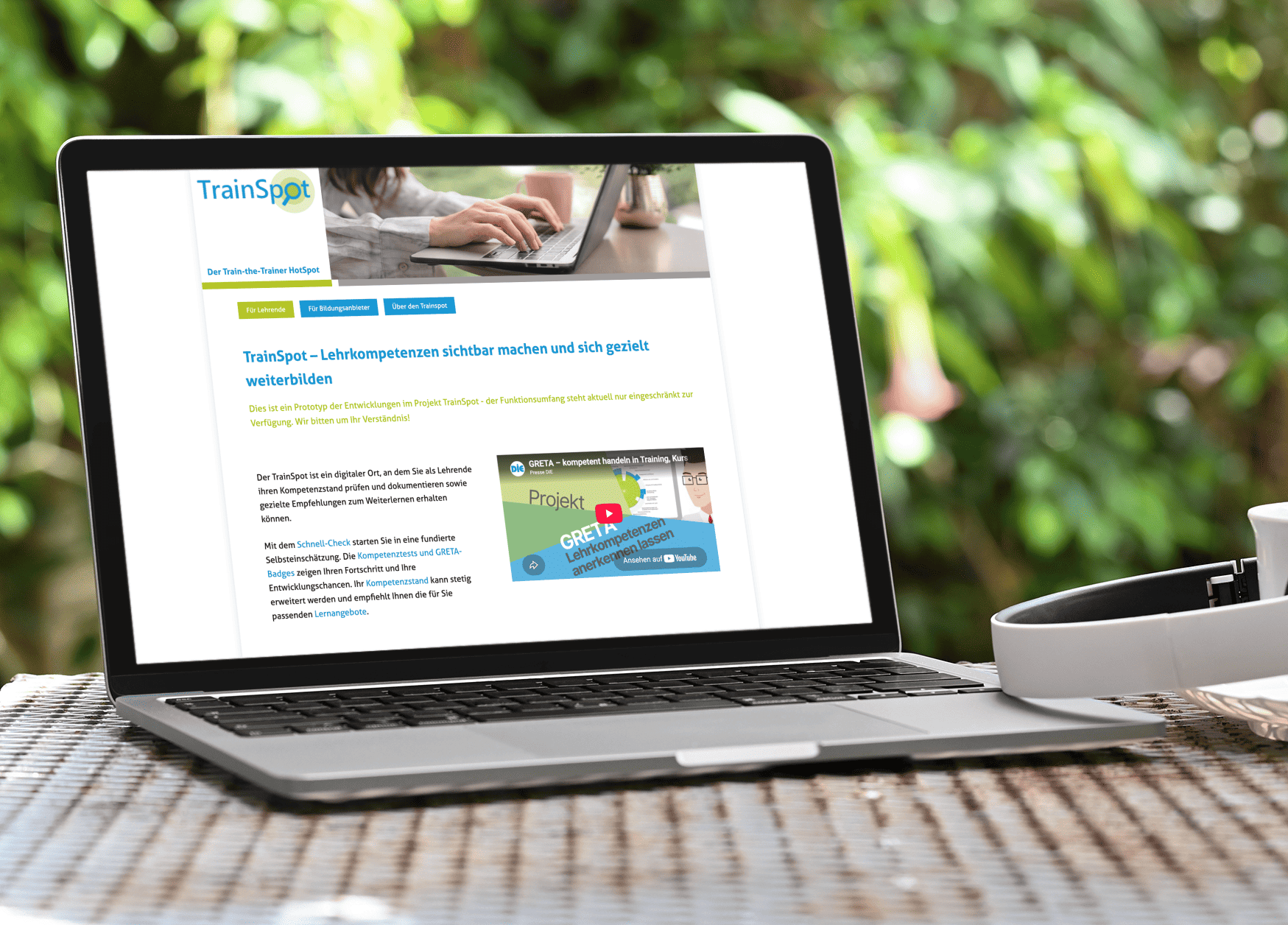 KI generiert: Das Bild zeigt einen Laptop mit einer geöffneten Webseite des Projekts "TrainSpot", die sich auf die Sichtbarmachung und gezielte Weiterbildung von Lehrkompetenzen konzentriert, auf einem Holztisch im Freien.