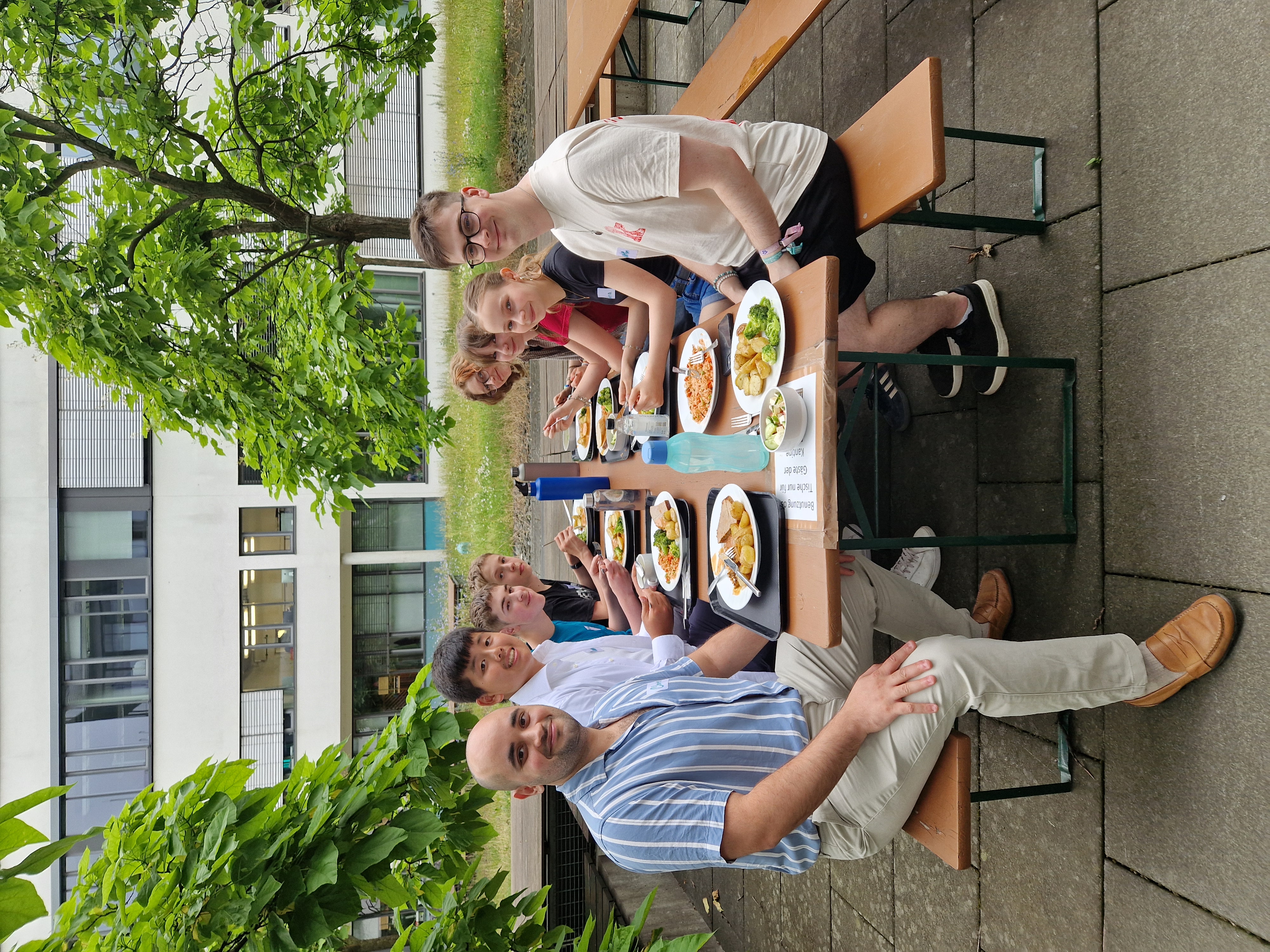 KI generiert: Das Bild zeigt eine Gruppe von Menschen, die gemeinsam an einem langen Holztisch im Freien sitzen und essen.