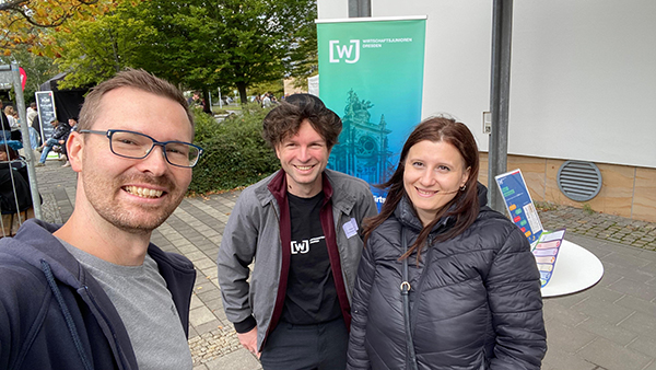 KI generiert: Auf dem Bild sind drei Personen, die lächelnd vor einem Infostand mit einem Banner posieren.